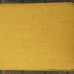 Ż&oacute;łta poduszka siedziska z widoczną fakturą materiału, leżąca na chodniku z szarej kostki brukowej, obok para szarych but&oacute;w sportowych.