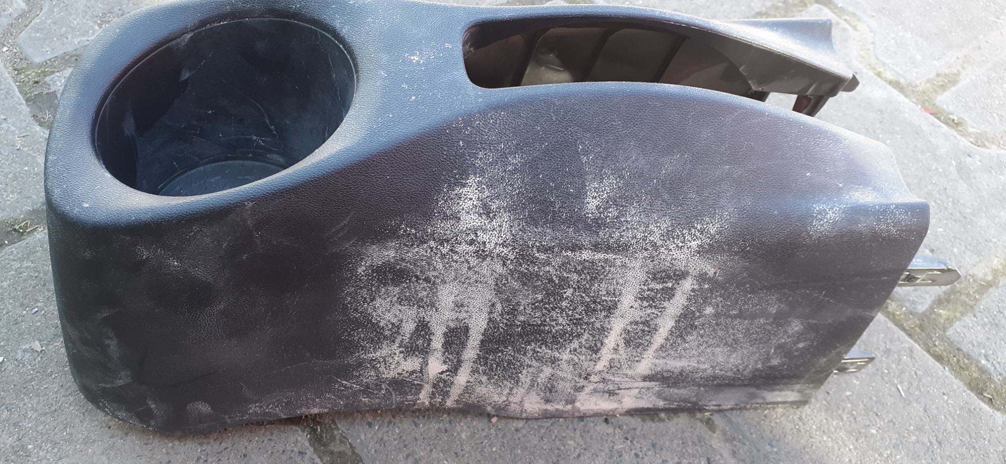 Czarny plastikowy element wnętrza samochodu z uchwytem na napoje, pokryty plamami i zabrudzeniami przed czyszczeniem, leżący na betonowej powierzchni.
