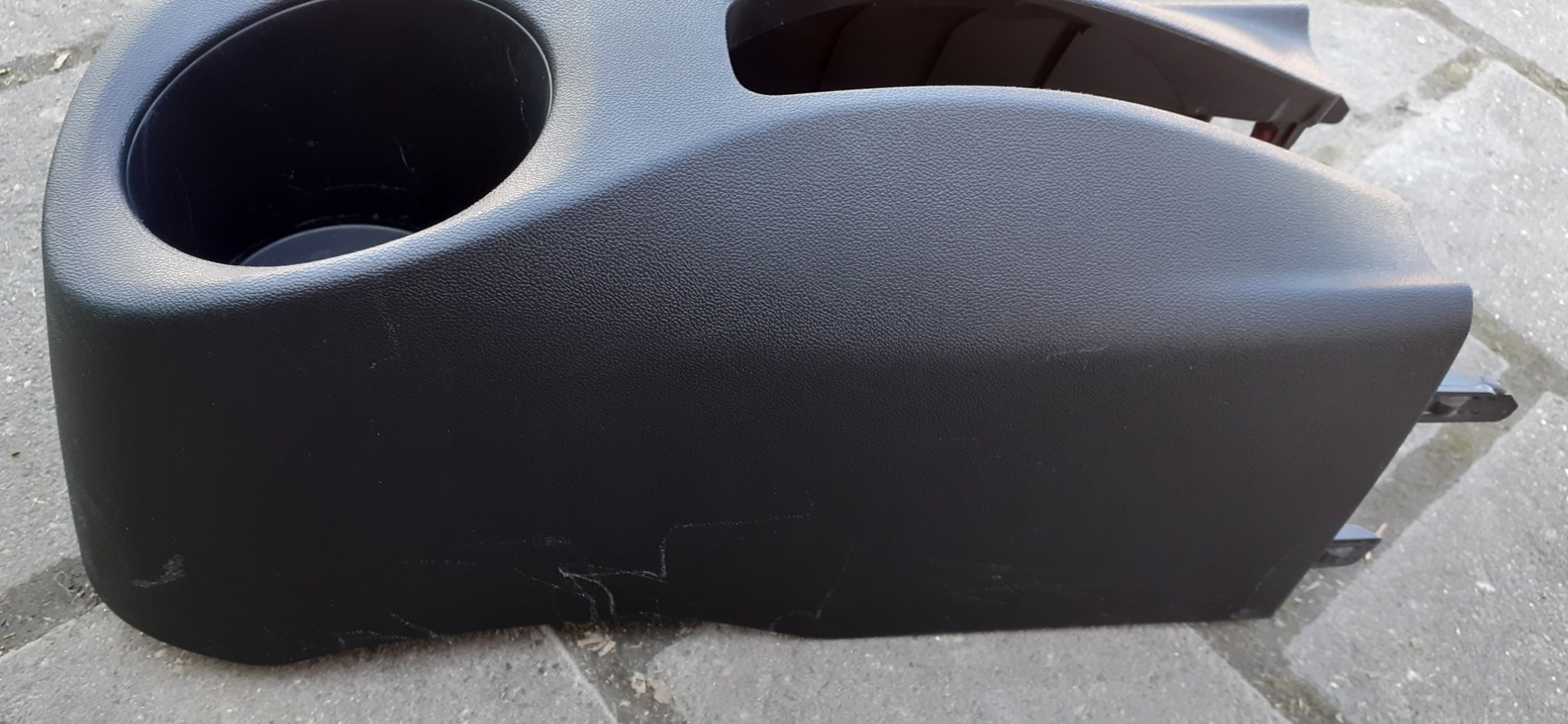 Czarny plastikowy element wnętrza samochodu z uchwytem na napoje, widoczne zarysowania i ślady użytkowania.