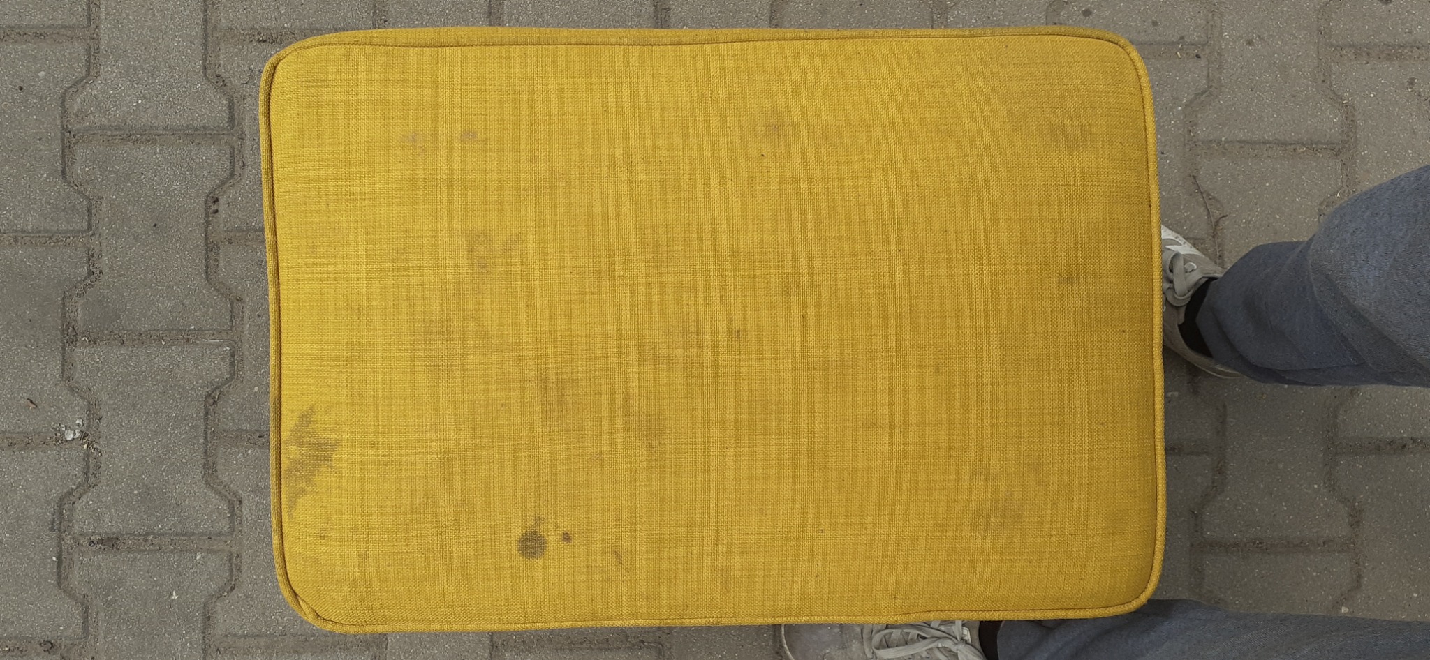 Żółty, kwadratowy podnóżek tapicerowany z widocznymi plamami na tkaninie, sfotografowany z góry na tle szarej kostki brukowej, obok widoczne fragmenty butów i spodni osoby fotografującej. Stan...