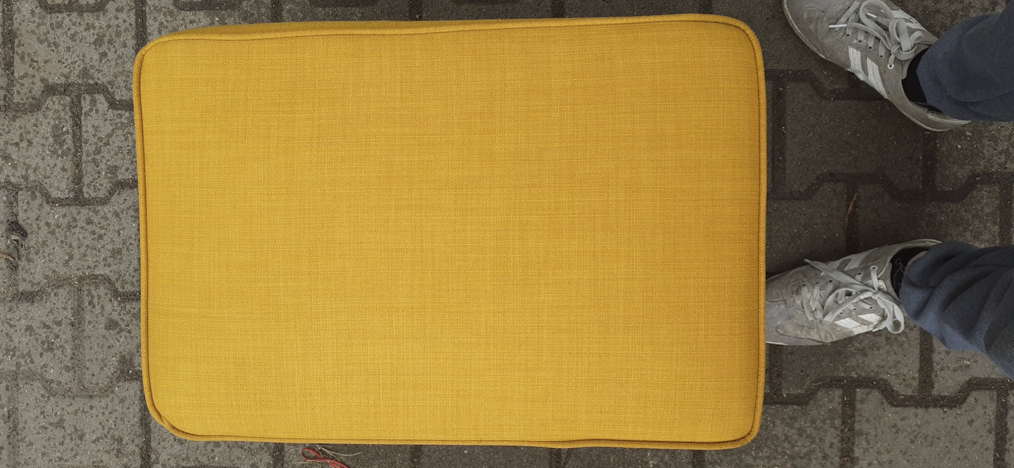 Żółta poduszka siedziska z widoczną fakturą materiału, leżąca na chodniku z szarej kostki brukowej, obok para szarych butów sportowych.