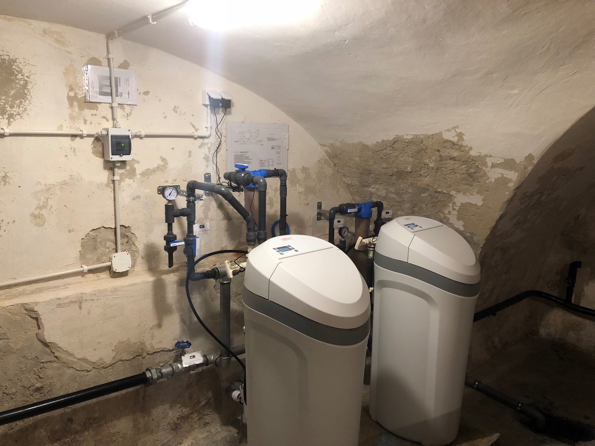 Dwa białe zmiękczacze wody Viessmann w piwnicy z łukowym sufitem i instalacją wodną, widoczne rury, zawory i manometr.