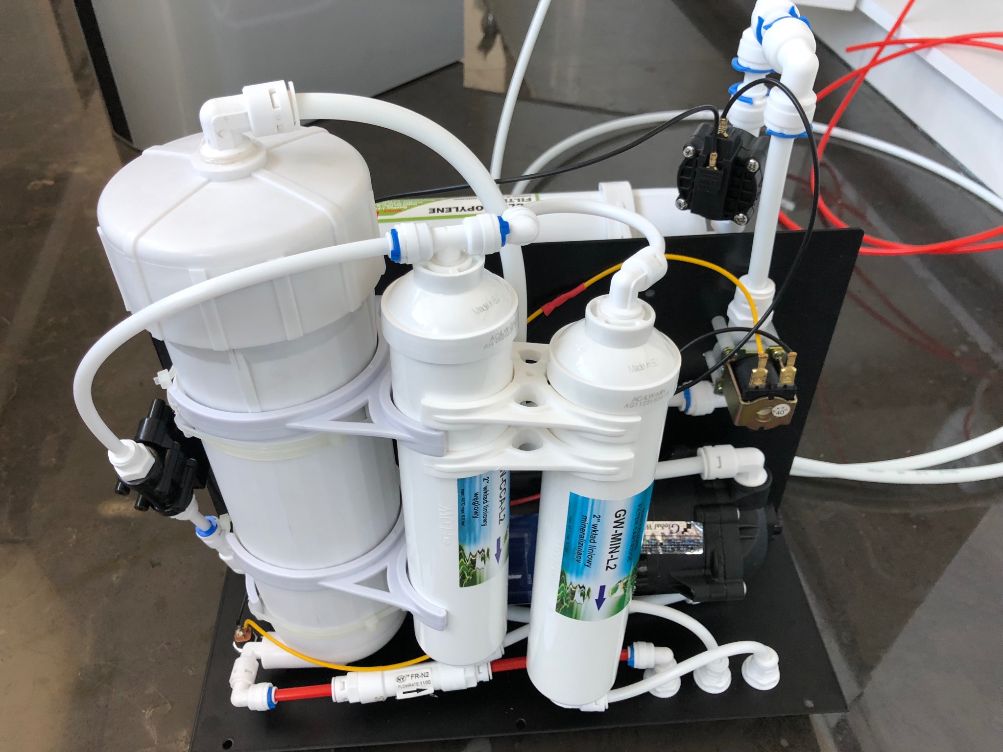 Zestaw filtrów do wody z widocznymi elementami: zbiornik, wkłady liniowe mineralizujące i węglowe, czarna rama montażowa oraz białe i kolorowe przewody.