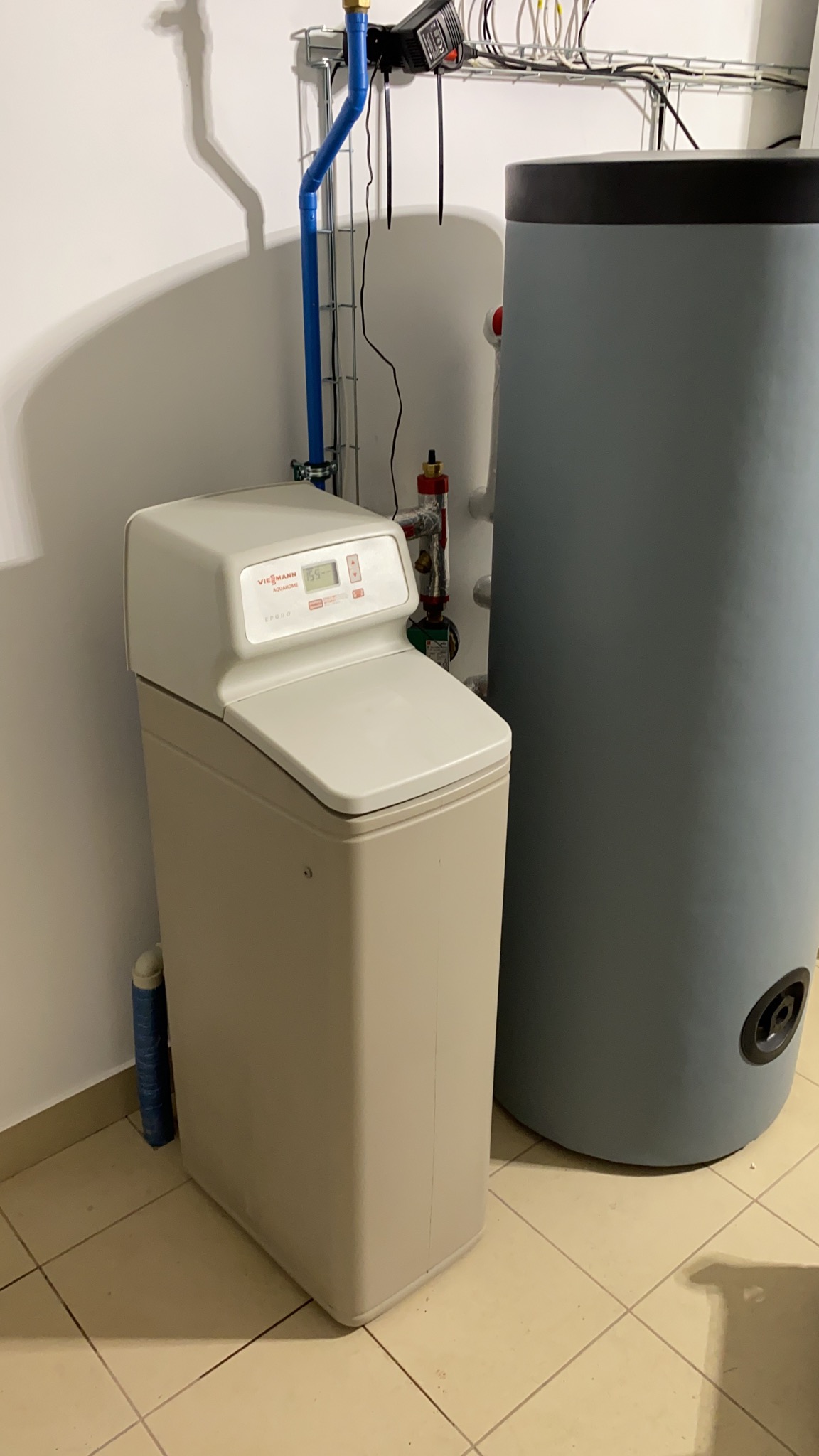 Beżowy zmiękczacz wody Viessmann Aquahome obok szarego zbiornika buforowego, widoczne rury instalacji wodnej i elektrycznej na tle białej ściany i beżowej podłogi z płytek.