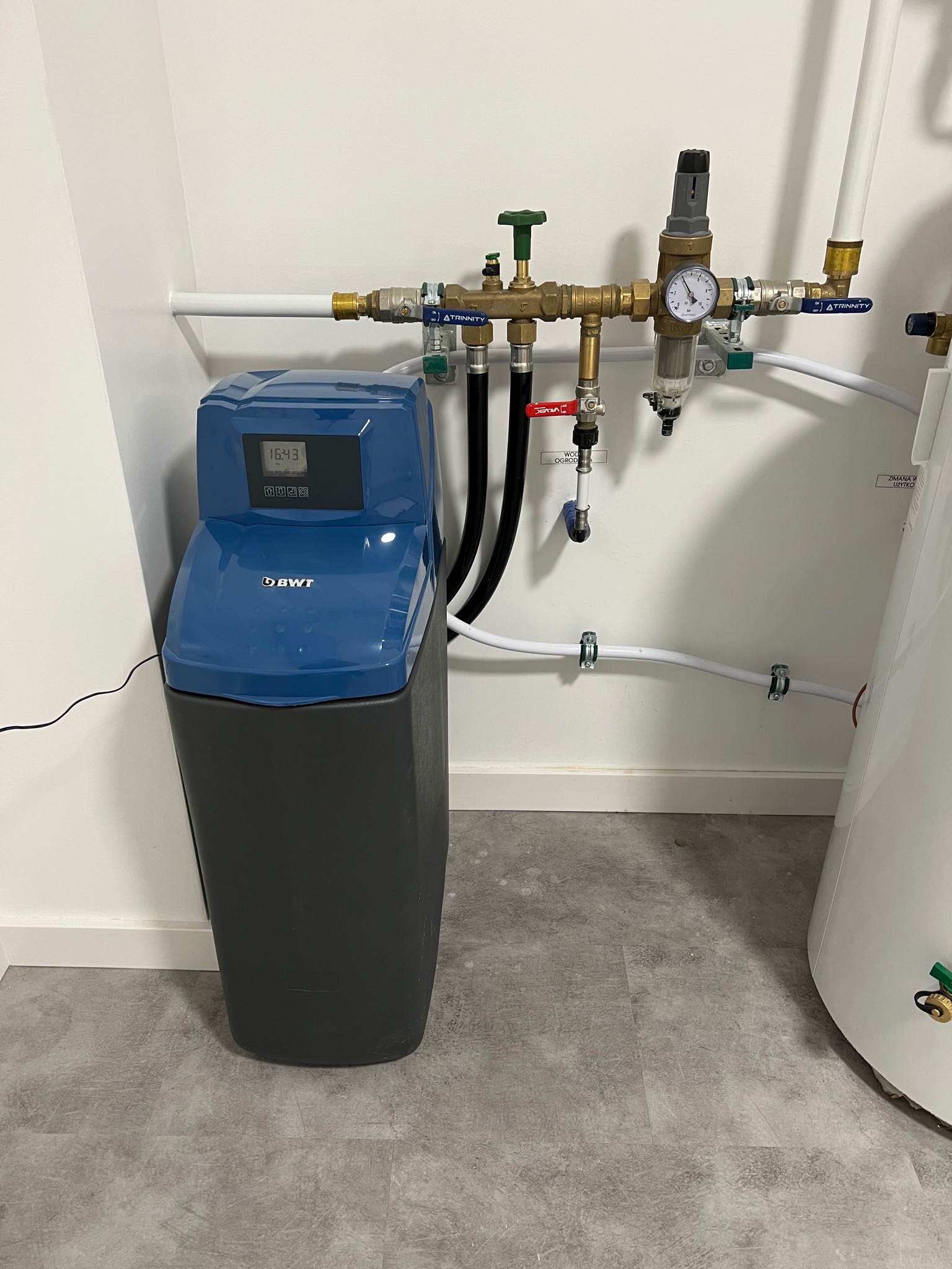 Zmiękczacz wody BWT zainstalowany w pomieszczeniu technicznym, widoczne rury miedziane i plastikowe, manometr oraz elementy instalacji wodnej. Na wyświetlaczu urządzenia widoczny aktualny czas.
