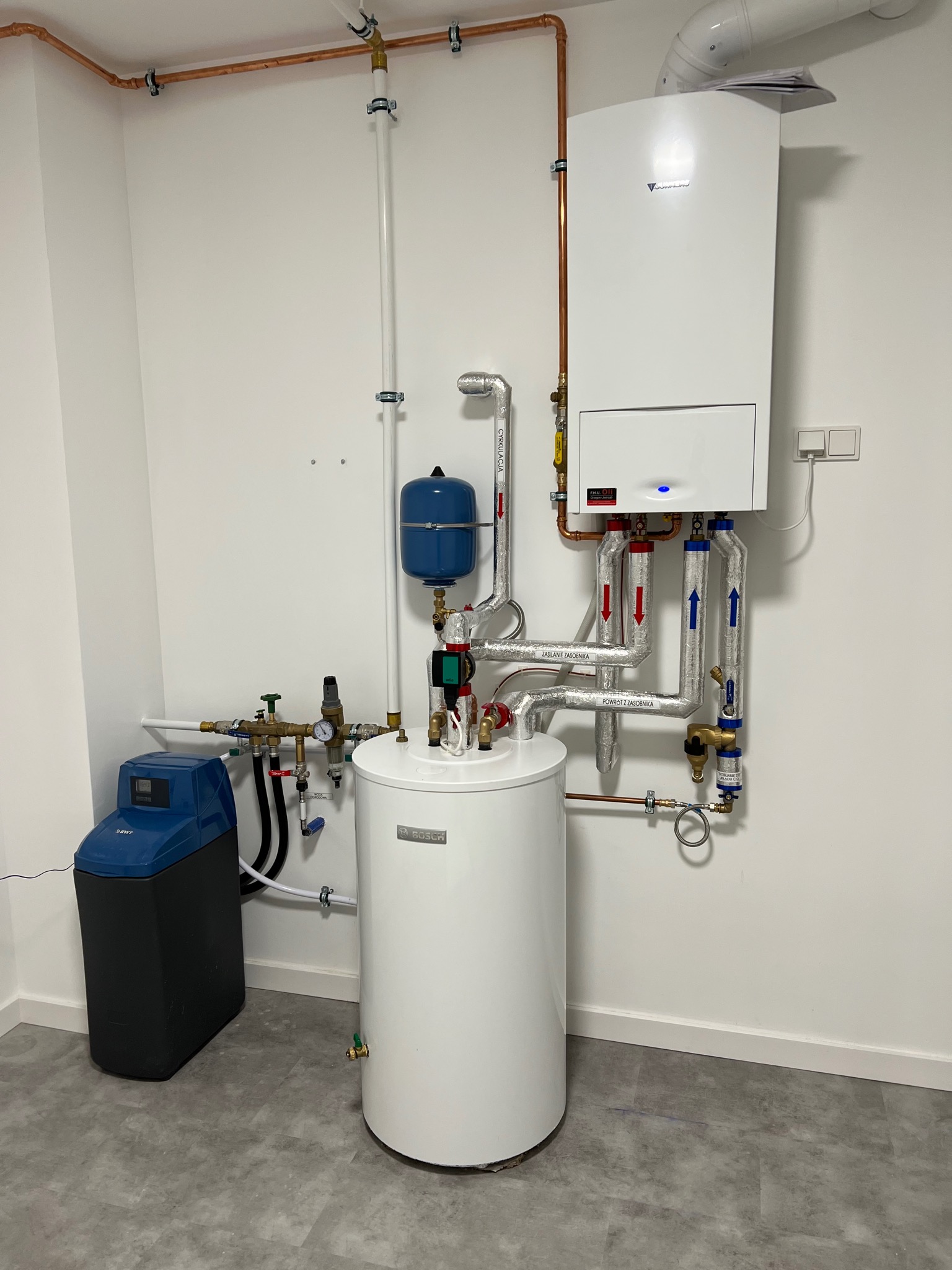 Biały kondensacyjny kocioł grzewczy Viessmann z podłączonym zasobnikiem ciepłej wody Bosch, widoczne rury miedziane i stalowe z izolacją, oraz zmiękczacz wody BWT, na tle białej ściany i szarej...