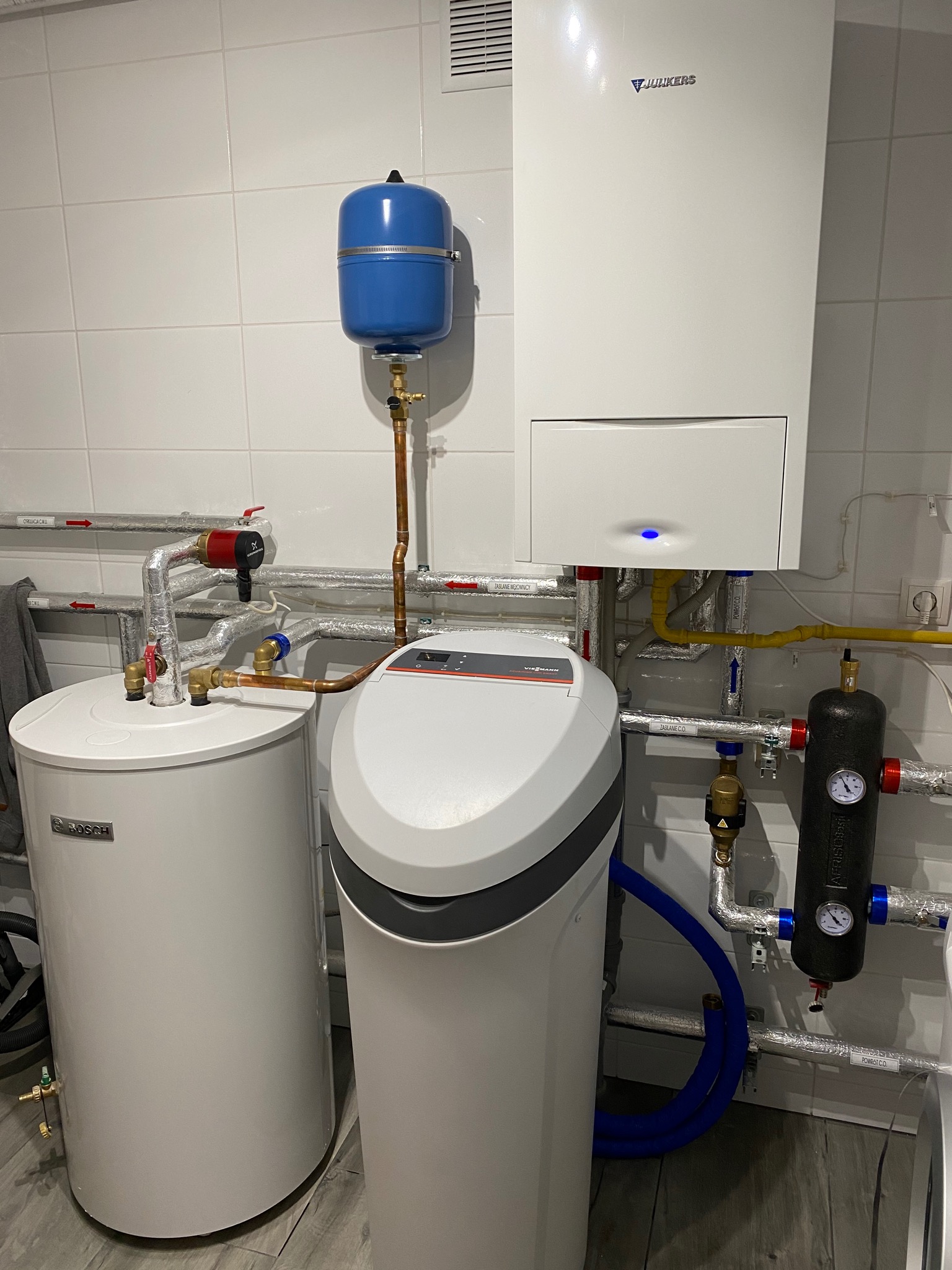 Kotłownia z białym kotłem Junkers, zasobnikiem Bosch, niebieskim naczyniem przeponowym i zmiękczaczem wody. Instalacja z rur miedzianych i stalowych z izolacją, manometry na czarnym elemencie.