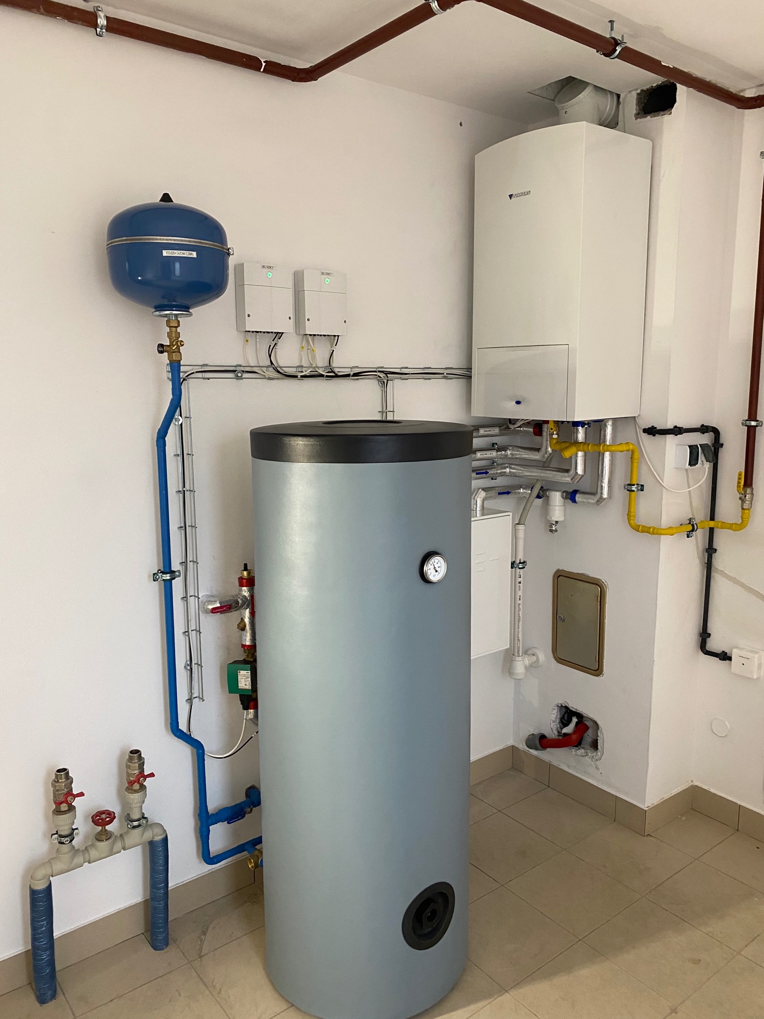 Kocioł gazowy Junkers wiszący na ścianie, obok pionowy, szary zasobnik ciepłej wody z manometrem, niebieski zbiornik wyrównawczy i rury w kolorze miedzi, żółte rury gazowe, białe ściany i beżowa...