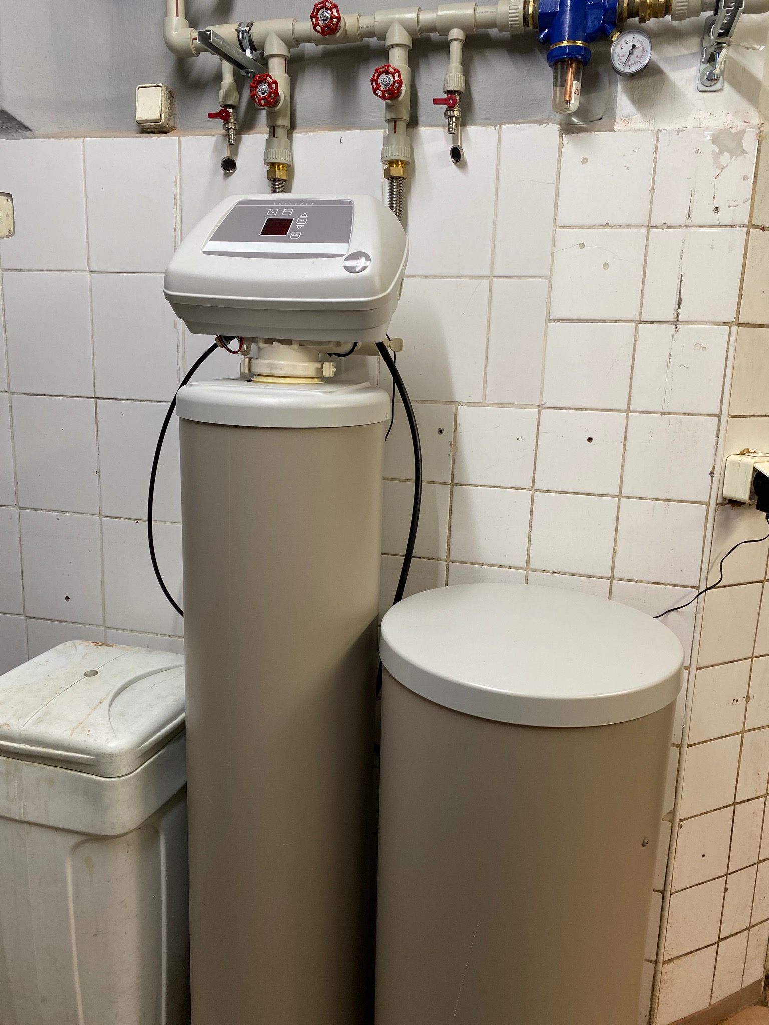 Centralny zmiękczacz wody z elektronicznym sterowaniem, zainstalowany w pomieszczeniu z białymi, kwadratowymi płytkami, widoczne rury i zawory na ścianie.