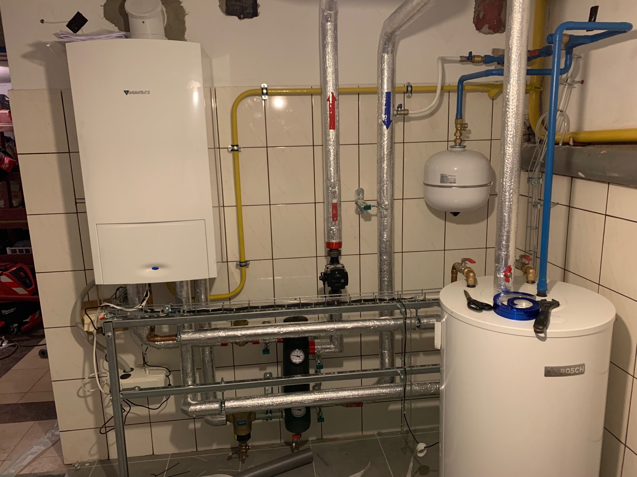 Biały kondensacyjny piec gazowy Junkers zamontowany na ścianie z glazurą, obok biały zasobnik ciepłej wody Bosch, rury instalacji grzewczej z zaworami i filtrami, piwnica.