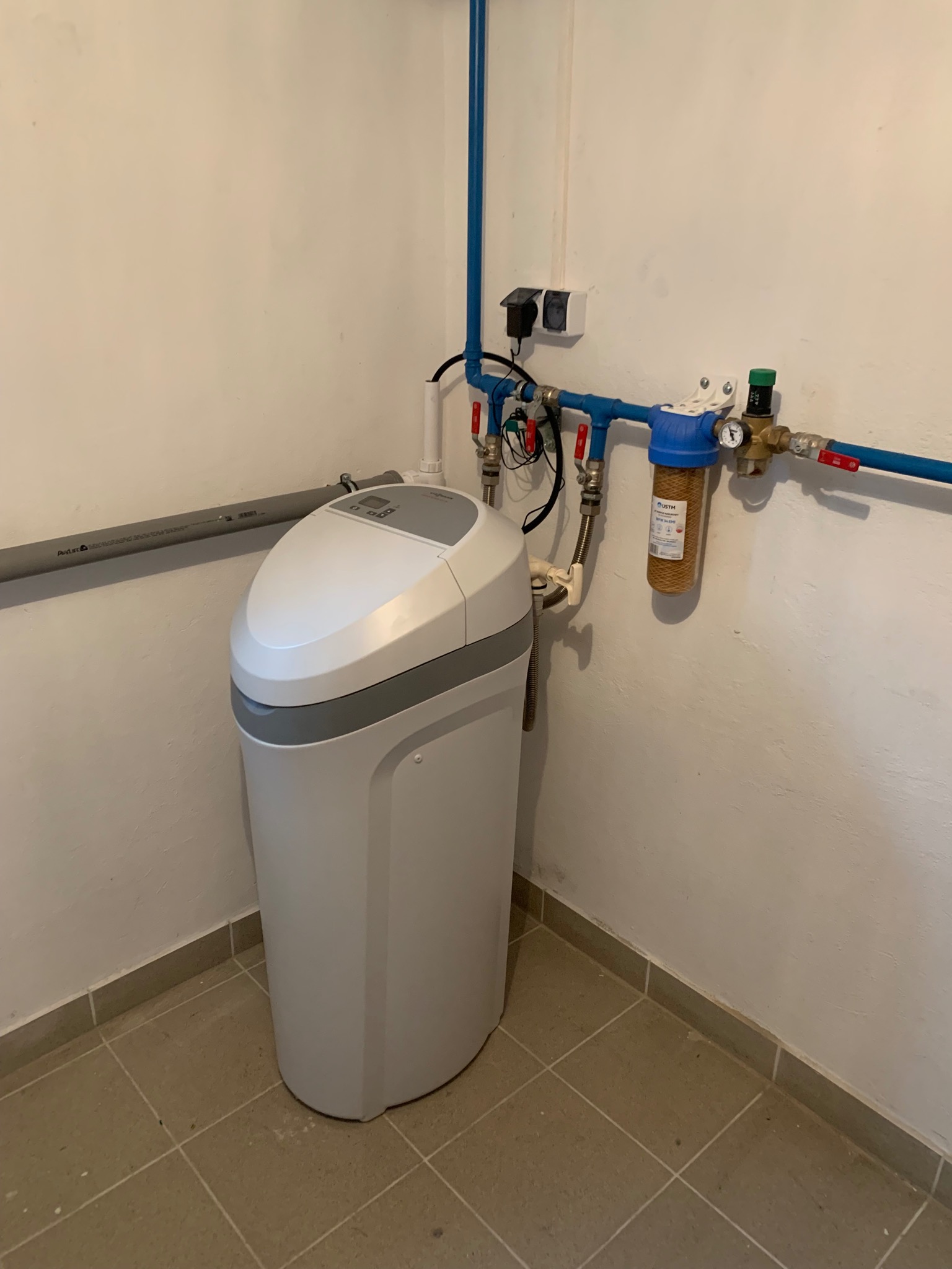 Biały zmiękczacz wody z filtrem mechanicznym, podłączony do niebieskich rur w pomieszczeniu z beżowymi płytkami podłogowymi.