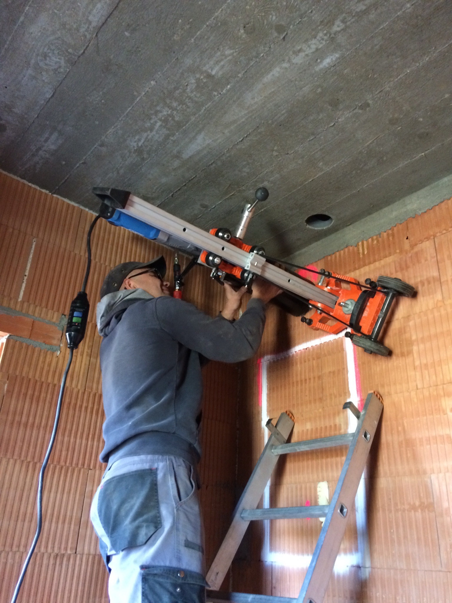 Pracownik w okularach wierci otwór w betonowym suficie za pomocą wiertnicy diamentowej, stojąc na drabinie w pomieszczeniu z ceglanymi ścianami.