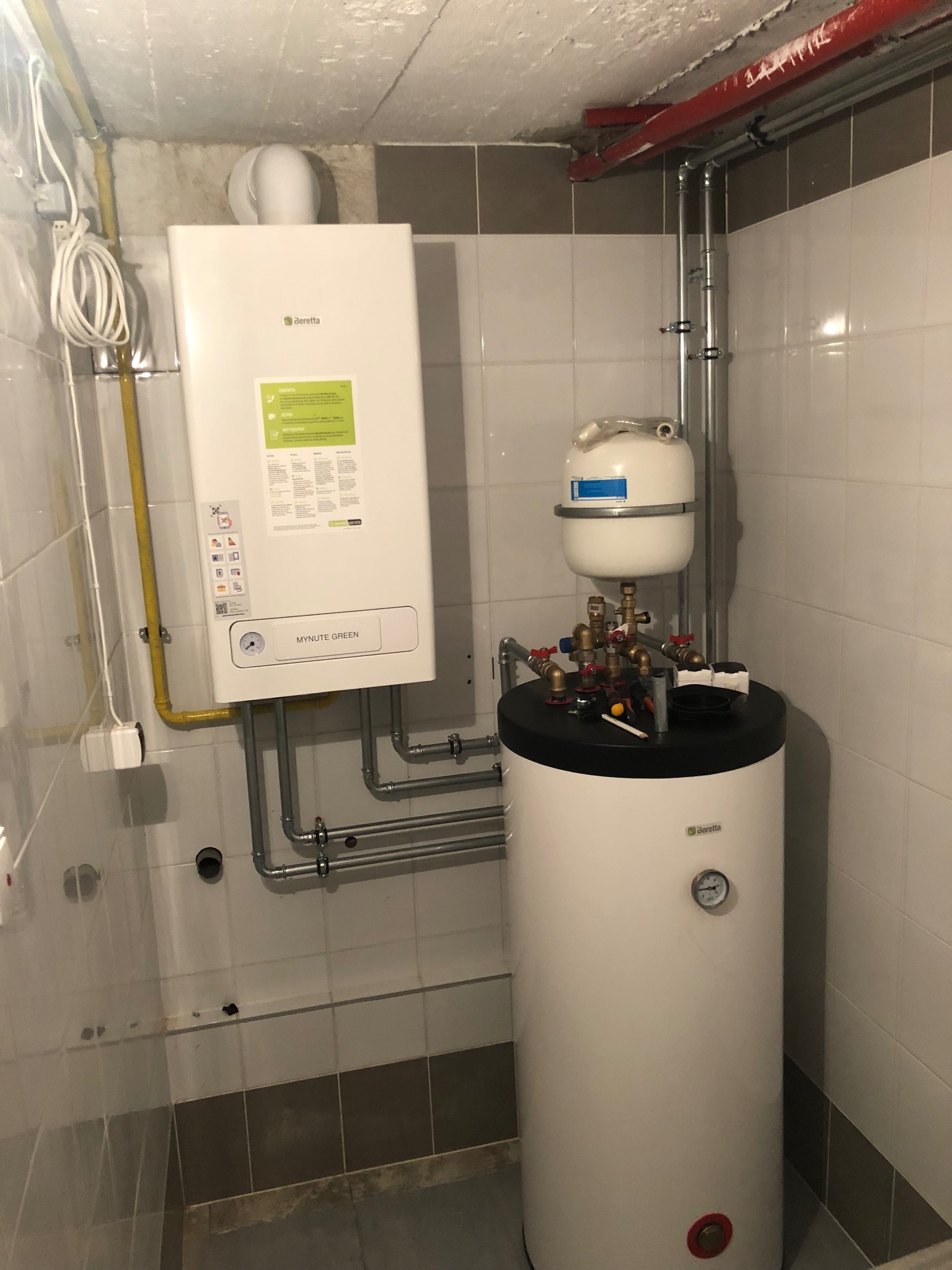 Biały, wiszący kocioł gazowy Beretta Mynute Green z podłączonymi rurami gazowymi i grzewczymi, obok biały zasobnik ciepłej wody użytkowej Beretta z manometrem, zamontowane w pomieszczeniu...