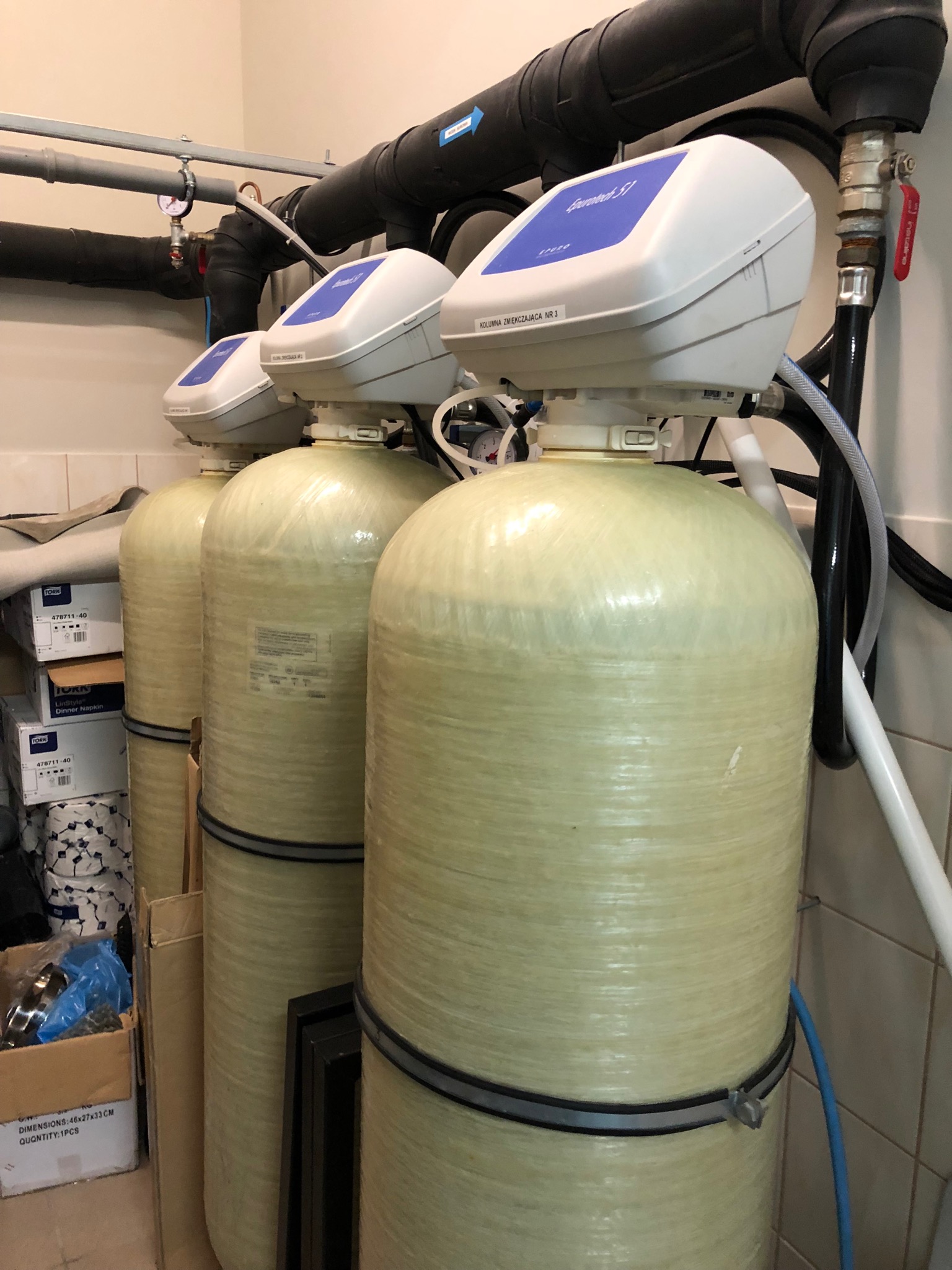 Trzy kolumny zmiękczające wodę Eurotech 51, połączone rurami, z widocznymi etykietami 'Kolumna zmiękczająca nr 3', w tle białe kafelki i kartony z napisem 'Tork'.