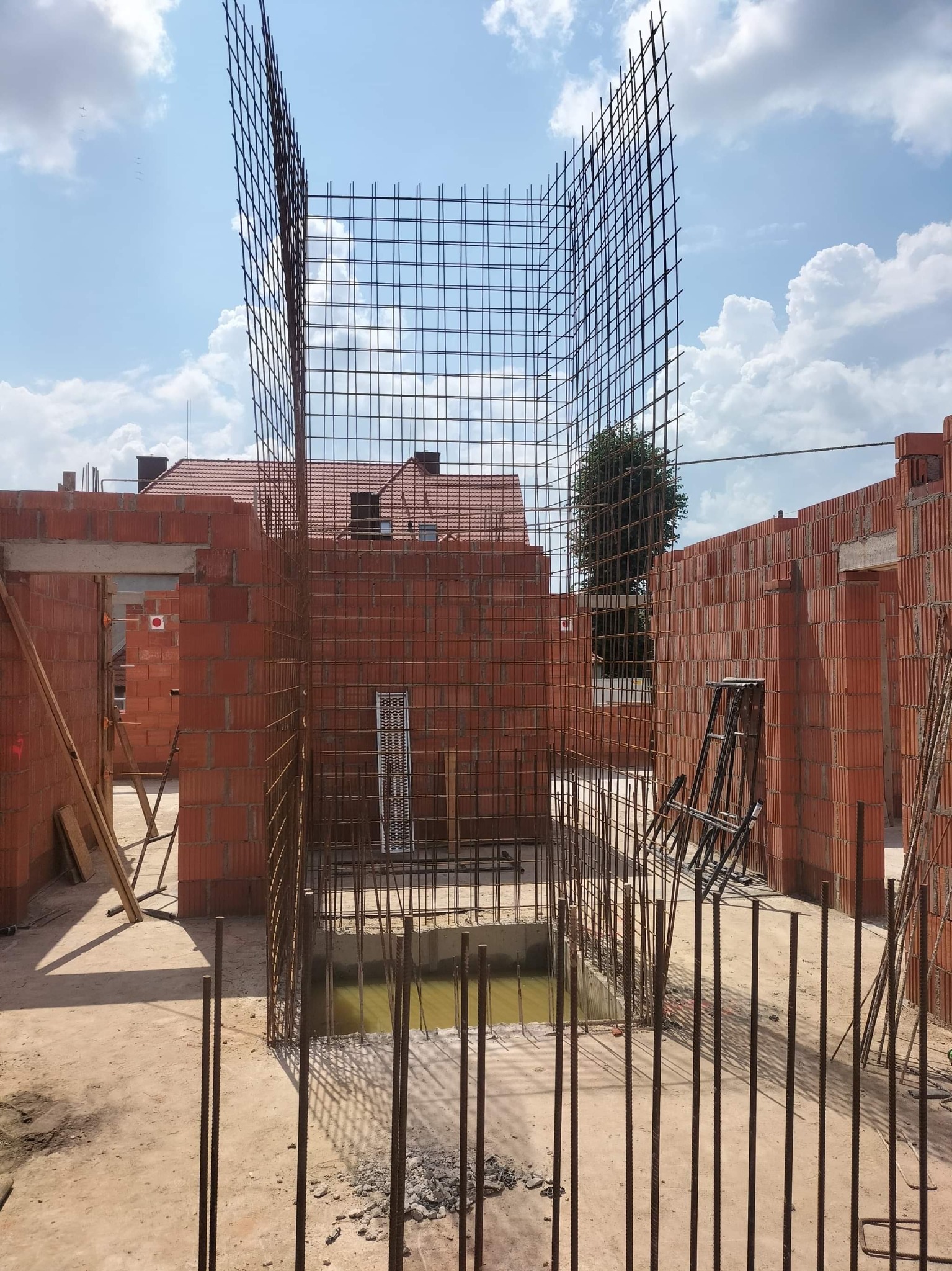 Zbrojenie pionowe w wykopie, przygotowane do zalania betonem, widoczne ściany z czerwonej cegły dziurawki i niebo z chmurami w tle.