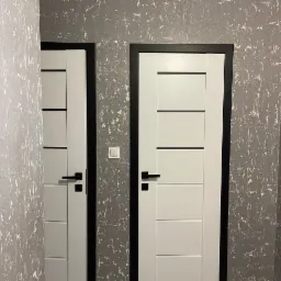 Dwa białe, nowoczesne drzwi wewnętrzne z czarnymi ościeżnicami i klamkami na tle ściany z szarą, strukturalną farbą. Widoczny włącznik światła.
