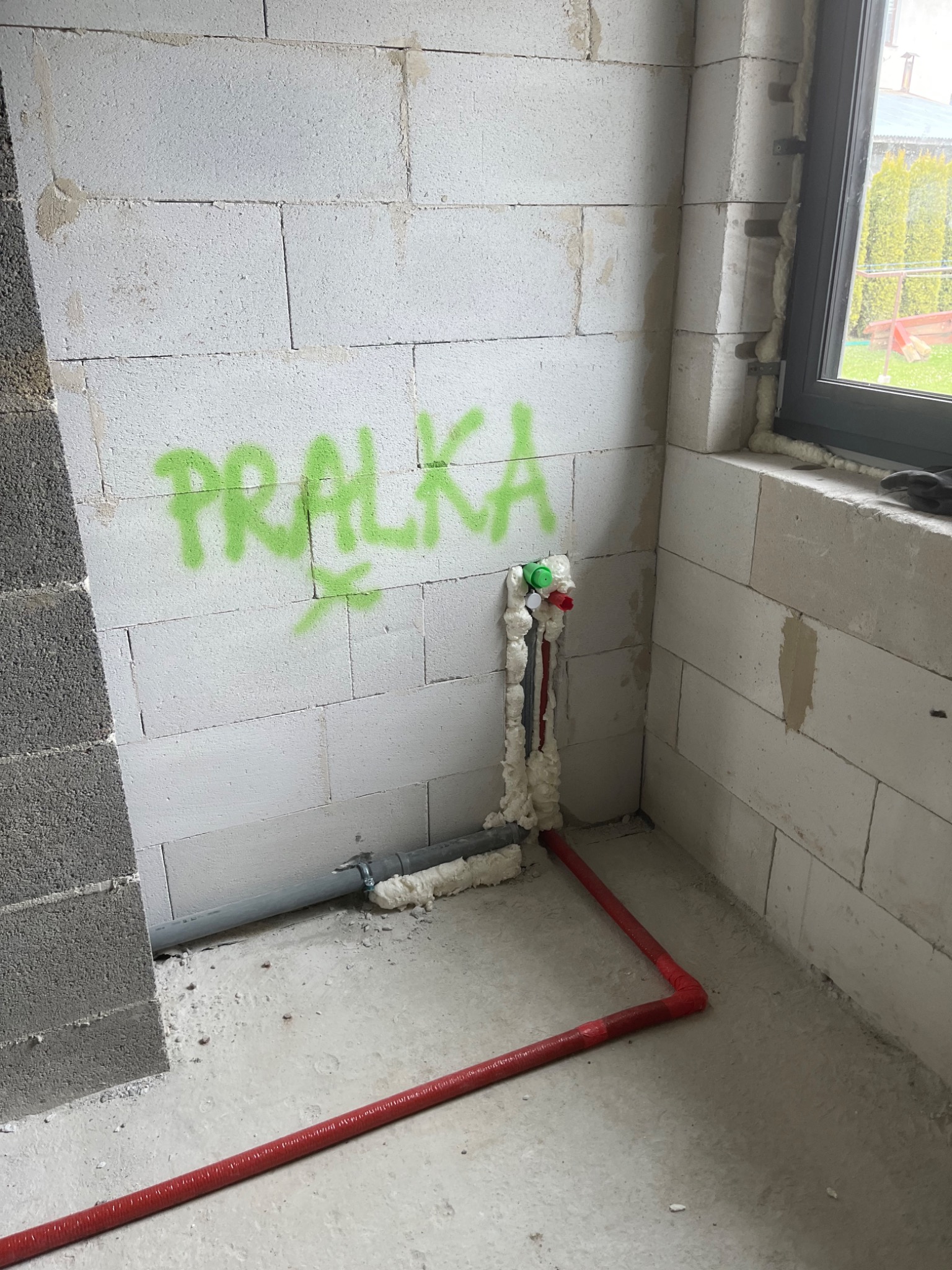 Wewnętrzne ściany z betonu komórkowego w trakcie budowy, z widocznymi rurami doprowadzającymi wodę, zabezpieczonymi pianką montażową i czerwoną rurą grzewczą, na ścianie napisane zielonym sprayem...