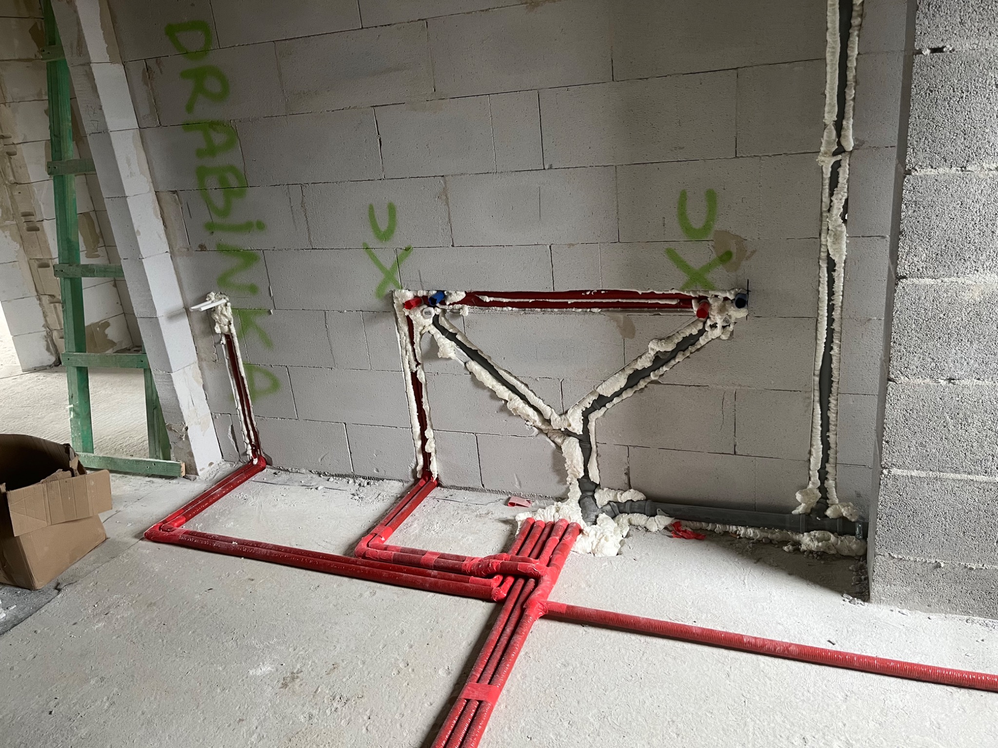 Surowy stan instalacji wodnej w budynku z bloków betonu komórkowego, widoczne czerwone rury ogrzewania podłogowego oraz rury kanalizacyjne zabezpieczone pianką montażową, na ścianie zielone napisy.