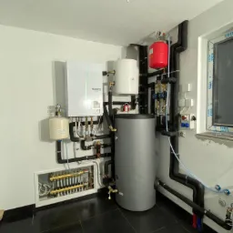 Kompletna instalacja grzewcza z szarą pionową jednostką zbiornika, białym modułem ściennym Ecodan z zielonym akcentem, czerwonym zbiornikiem wyrównawczym i licznymi rurami w czarnej izolacji...