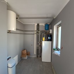 Lemon pompy ciepła - Zainstalowana pompa ciepła Daikin w szarej kotłowni w Toruniu. Widoczne rury, zbiornik wyrównawczy i zmiękczacz wody. Ujęcie z niskiego kąta.