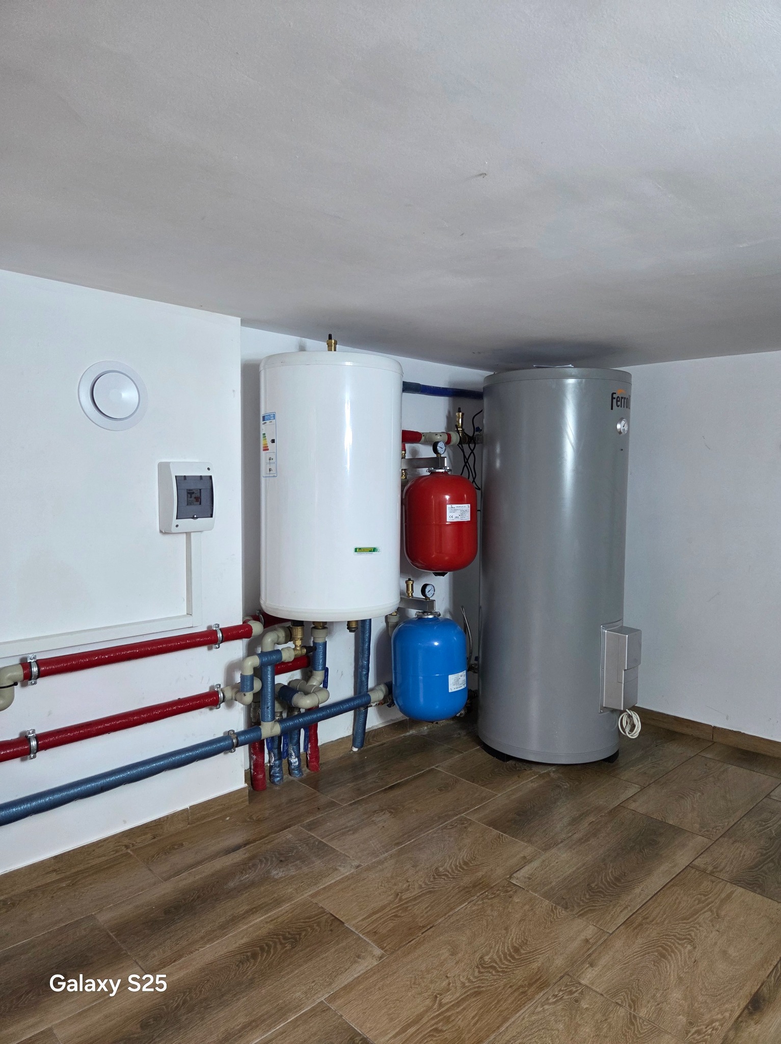 Zestaw zbiorników buforowych i instalacji grzewczej w kotłowni z brązowymi panelami podłogowymi. Widoczne rury w kolorach czerwonym i niebieskim.