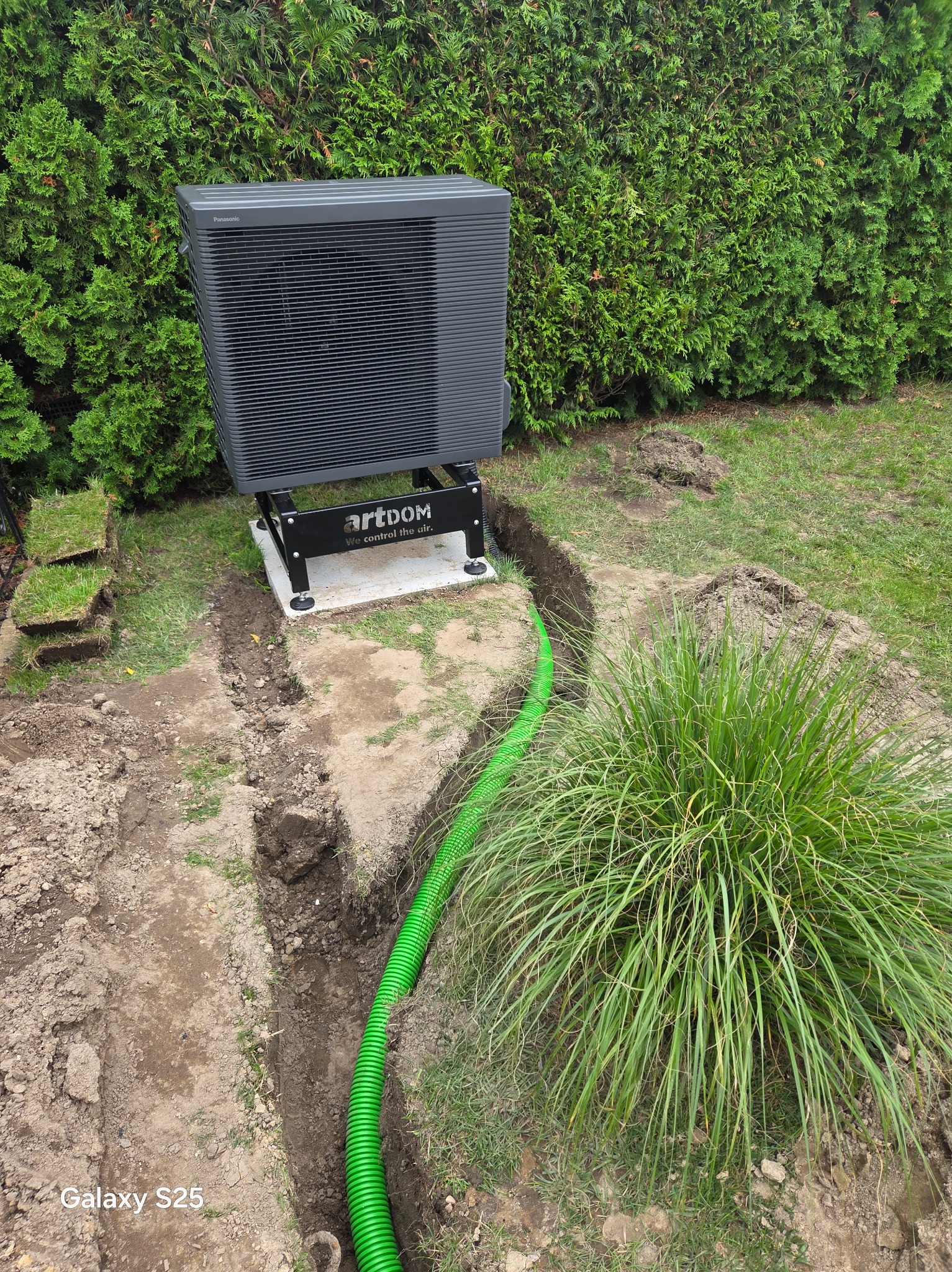 Montaż zewnętrznej jednostki pompy ciepła Panasonic na betonowej podstawie. W wykopie widoczna zielona rura. Trawa i tuje w tle.