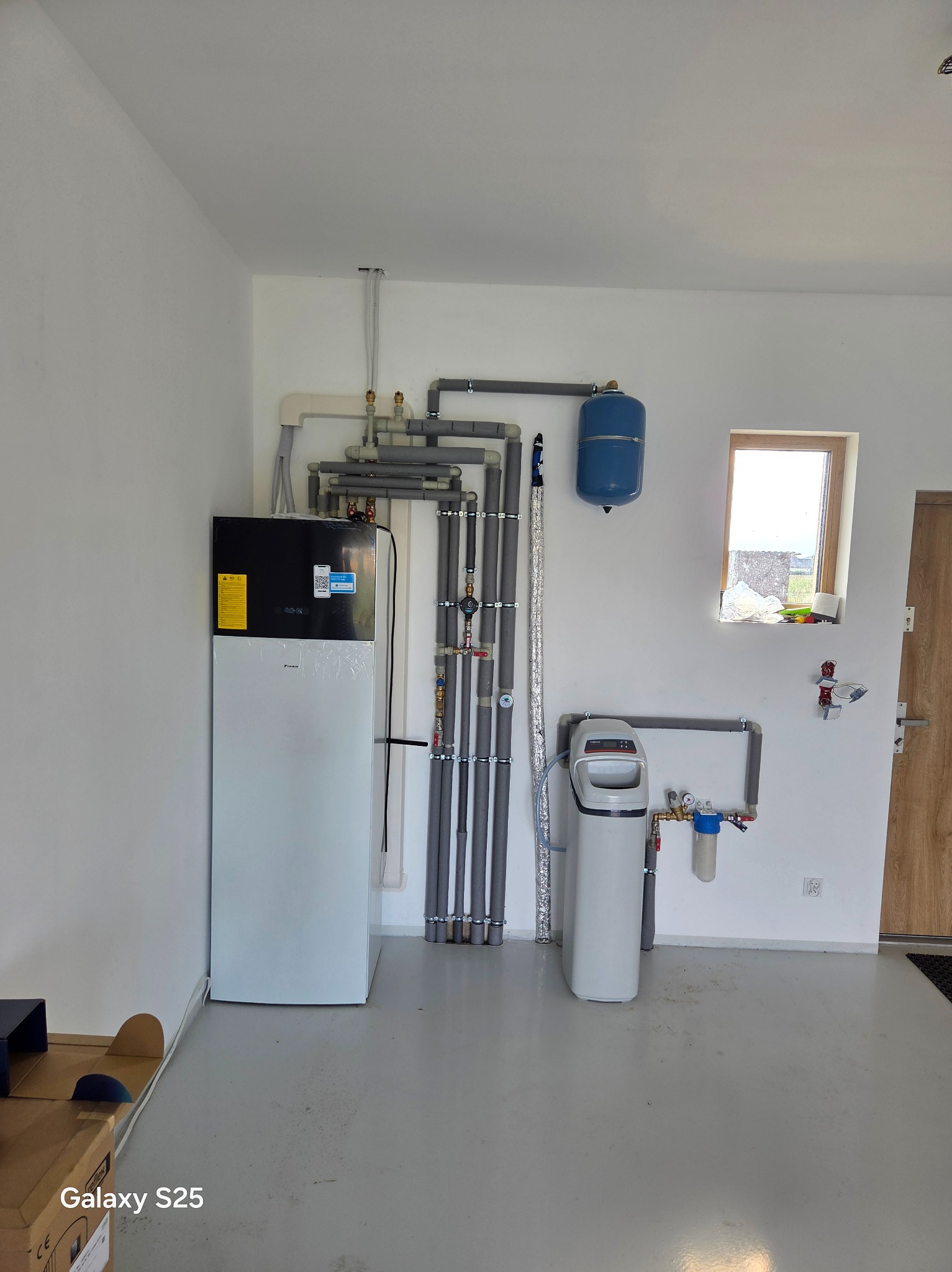 Zainstalowana pompa ciepła Daikin w jasnym pomieszczeniu z szarymi rurami, zbiornikiem wyrównawczym i filtrem wody. Widoczne okno i drzwi.