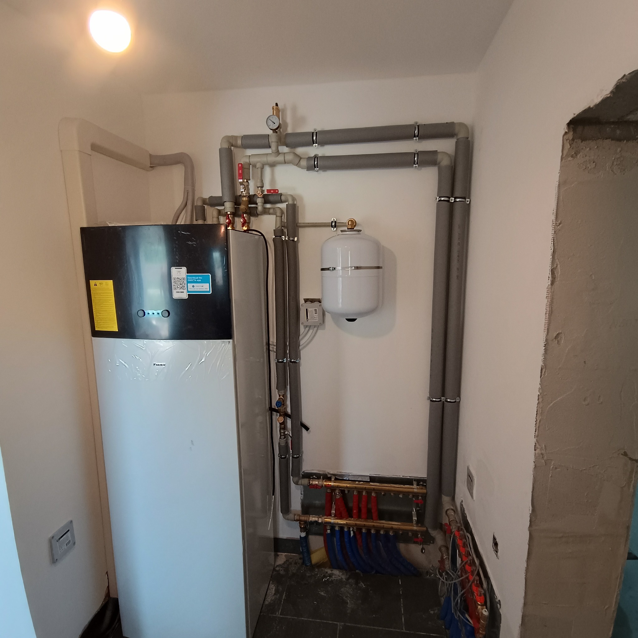 Zainstalowana pompa ciepła Daikin z widocznymi rurami instalacji grzewczej i zbiornikiem wyrównawczym w pomieszczeniu z surowym wykończeniem ściany.