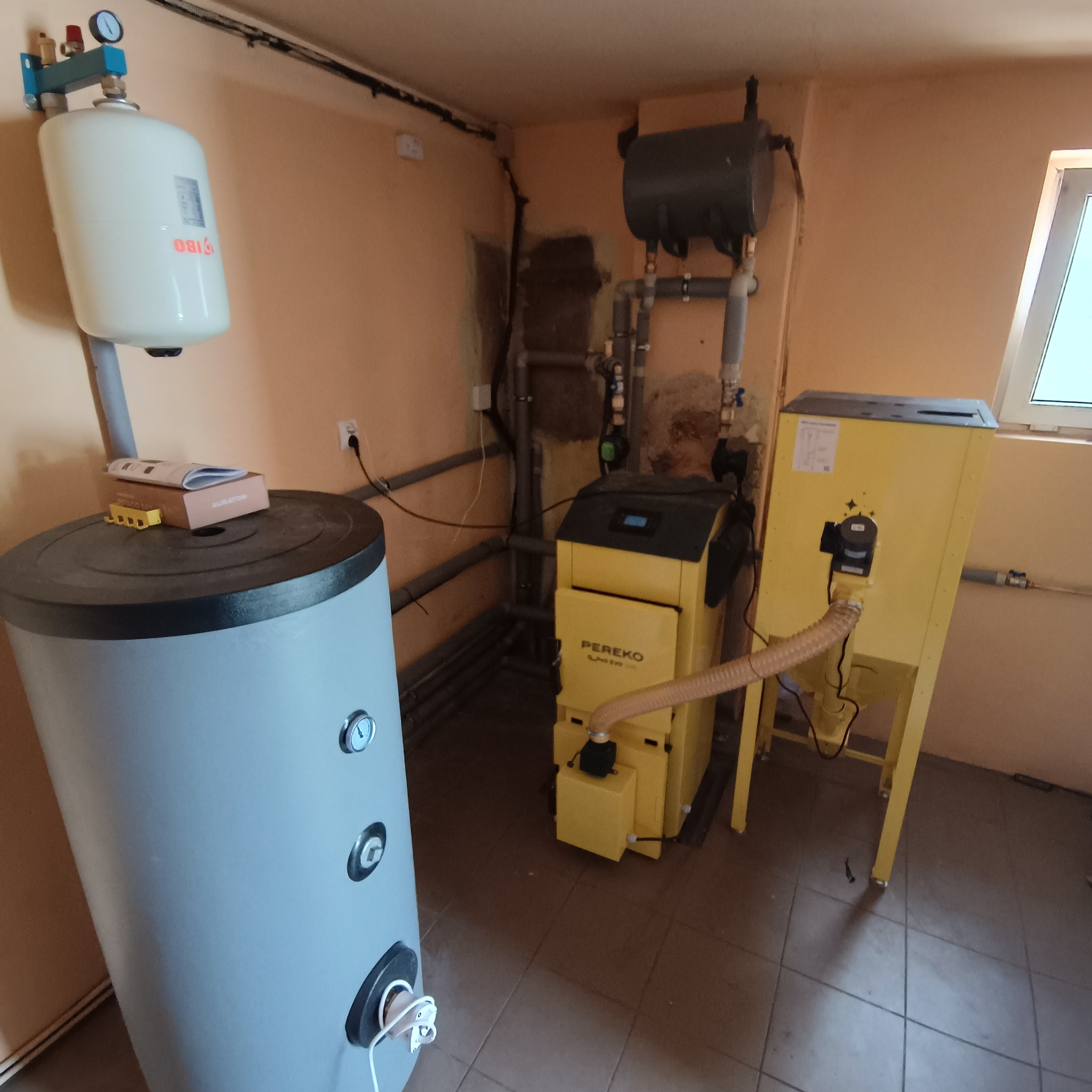 Żółty piec na paliwo stałe Pereko, szary zbiornik IBO, instalacja CO w kotłowni. Widoczne rury, zawory i osprzęt. Na zbiorniku leżą akcesoria Auraton.