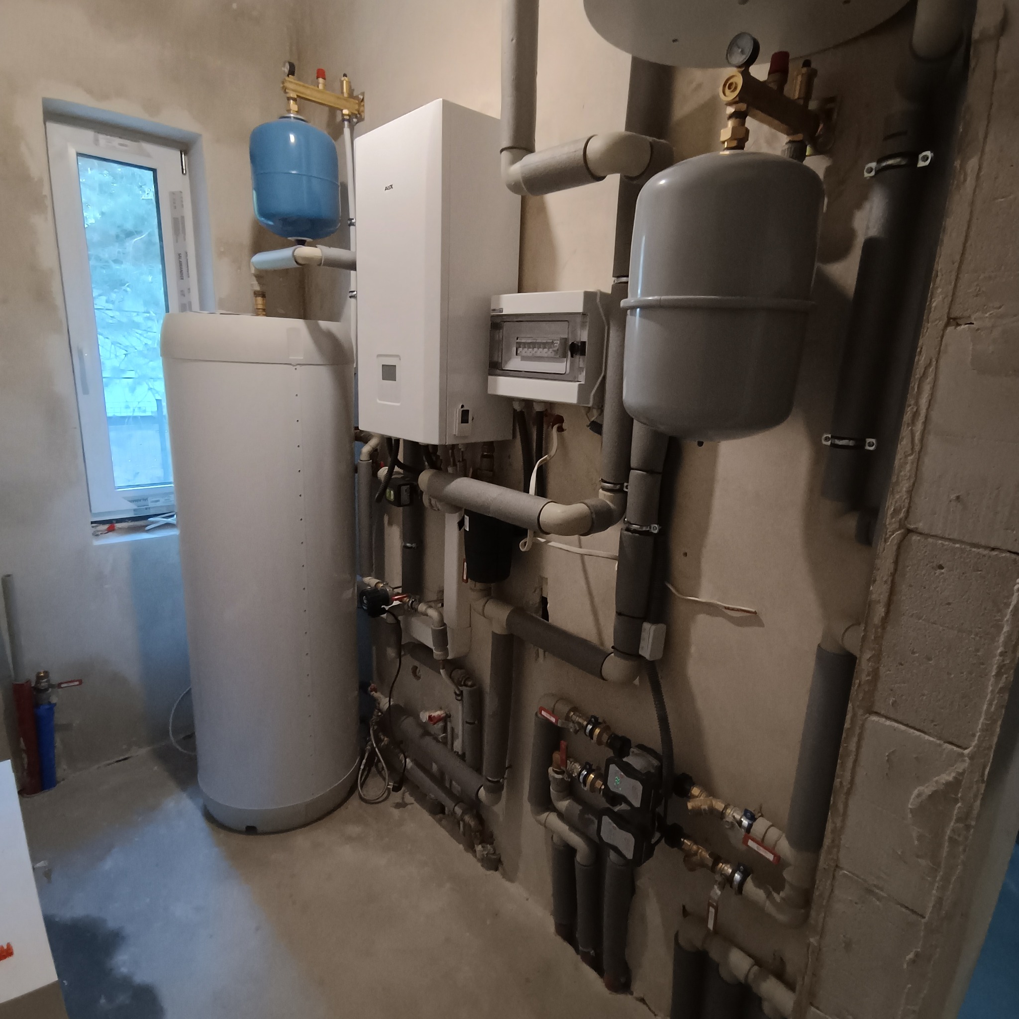 Złożona instalacja grzewcza z białym piecem, zbiornikiem i licznymi rurami w surowym pomieszczeniu z oknem. Widoczne elementy instalacji i przyłącza.