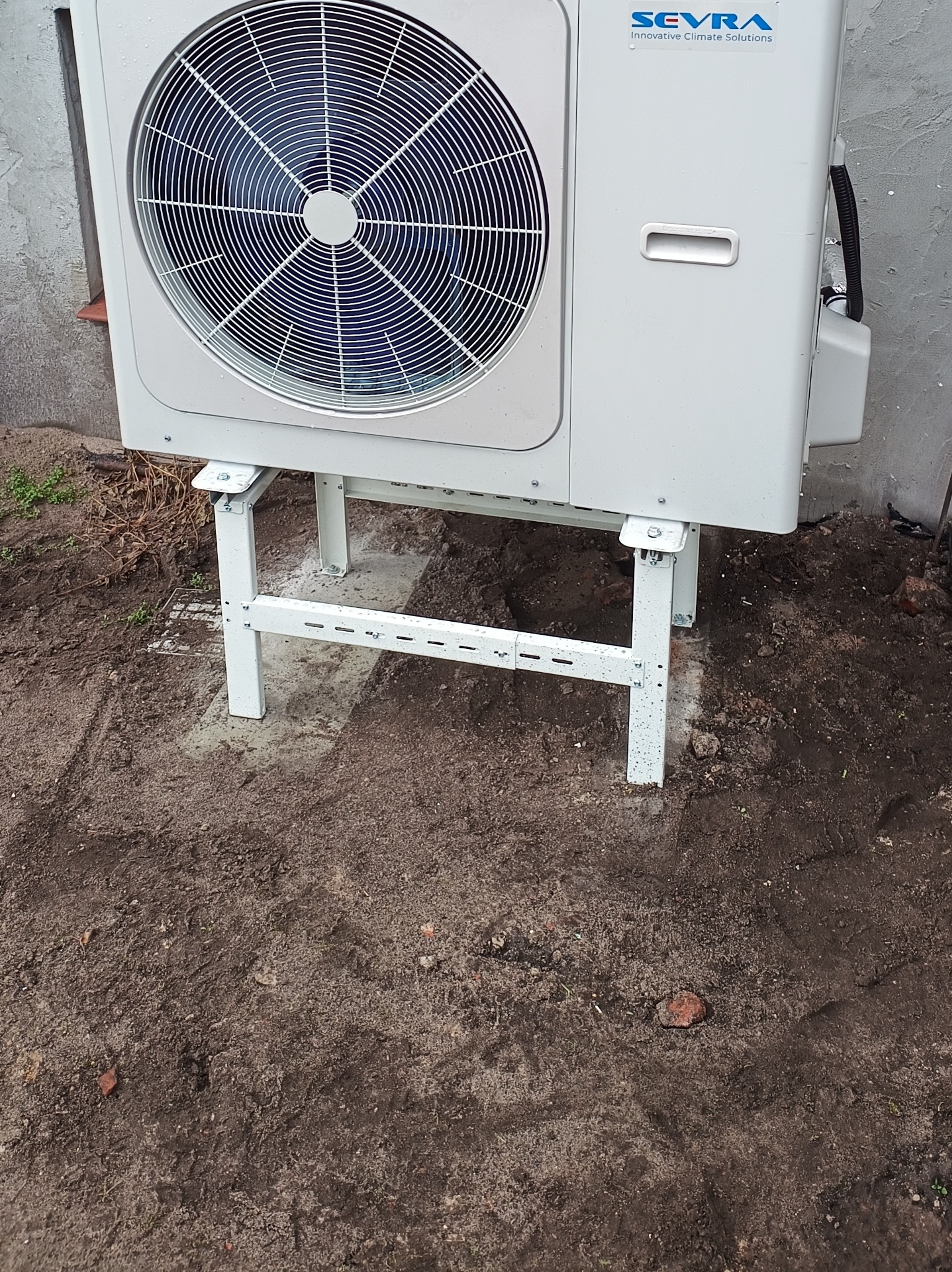 Biała pompa ciepła marki Sevra, zamontowana na metalowym stelażu, stoi na ziemi przy szarej ścianie. Widoczne detale montażu i otoczenie.