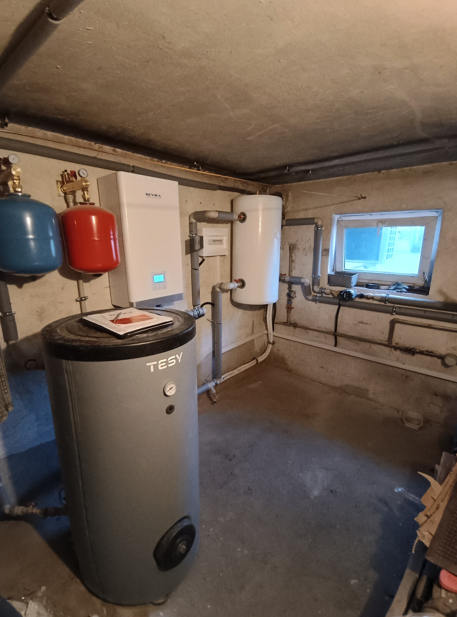 Instalacja kotła grzewczego Seyra i zasobnika TESY w piwnicy, z widocznymi rurami i elementami instalacji. Na zasobniku leży dokumentacja.