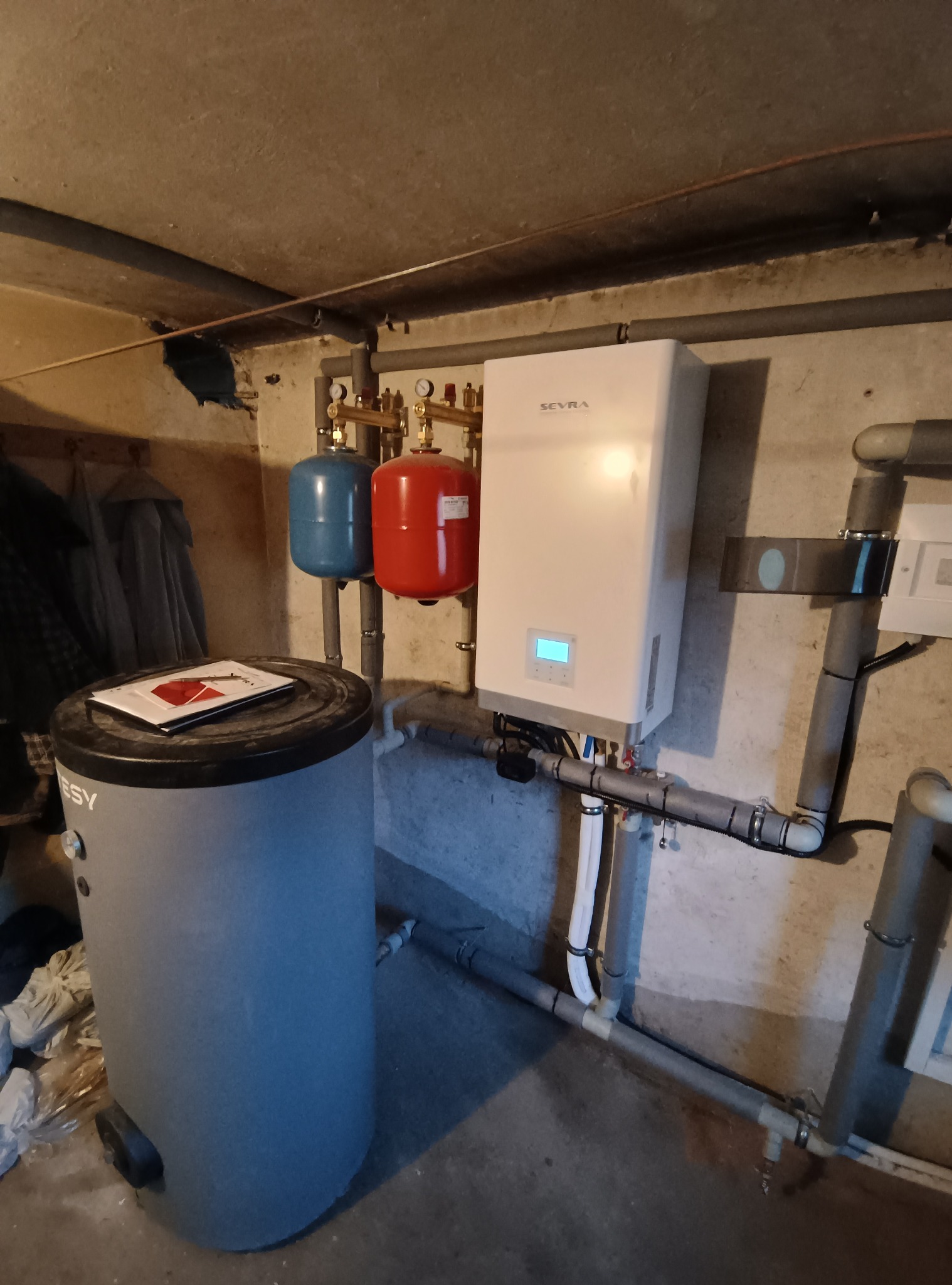 Zainstalowany biały piec gazowy SEVRA z czerwonym i niebieskim zbiornikiem wyrównawczym, szary zbiornik na wodę ESY i rury w piwnicy.