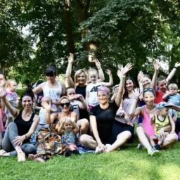 Grupa kobiet z dziećmi na trawie w parku, część z nich trzyma uniesione ręce, obok leży fioletowa mata do jogi.