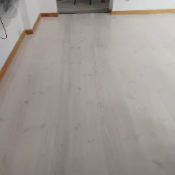 Jasne, bielone deski podłogowe z widocznym usłojeniem, świeżo położone w pokoju z drewnianymi listwami przypodłogowymi i drzwiami po lewej stronie.