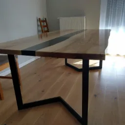 Stół z blatem z jasnego drewna z czarną żywicą pośrodku, na czarnych, metalowych nogach w kształcie trapezu, widoczne krzesła w tle.