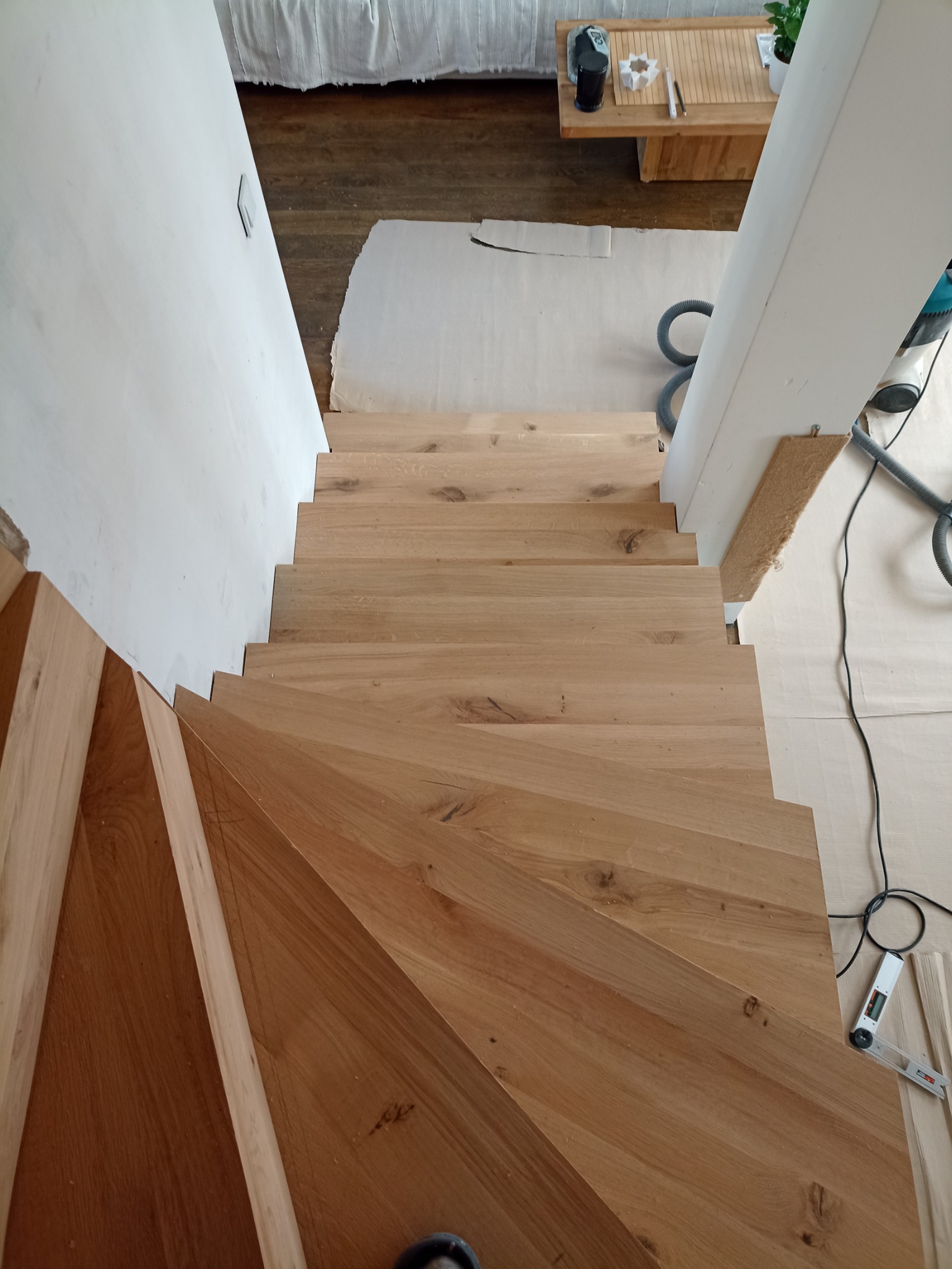 Drewniane schody z jasnego drewna dębowego, widok z góry w trakcie montażu, zabezpieczone podłogi i narzędzia stolarskie w tle.