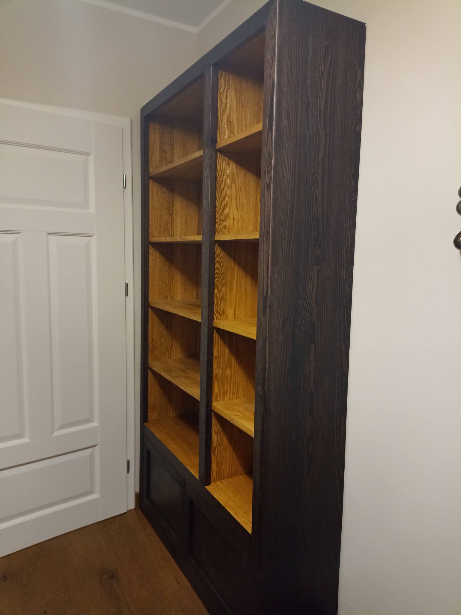 Wysoka, drewniana biblioteczka z ciemnobrązową ramą i jasnymi półkami, ustawiona przy białej ścianie i drzwiach, widoczna struktura drewna.