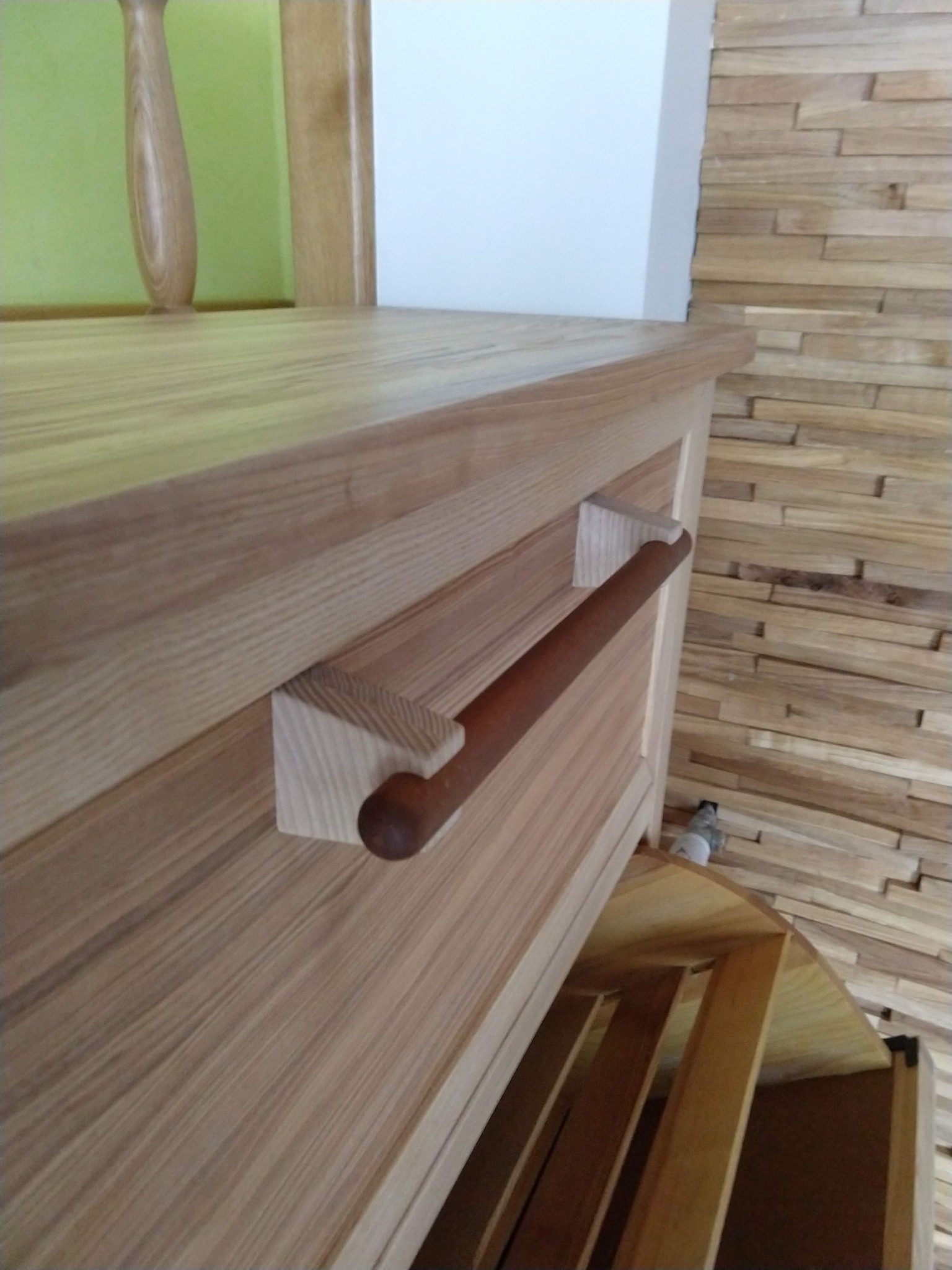 Fragment drewnianej szafy z wysuwanym relingiem na ręczniki, widoczny otwarty fragment szafki z półkami, ściana wyłożona drewnianymi panelami.