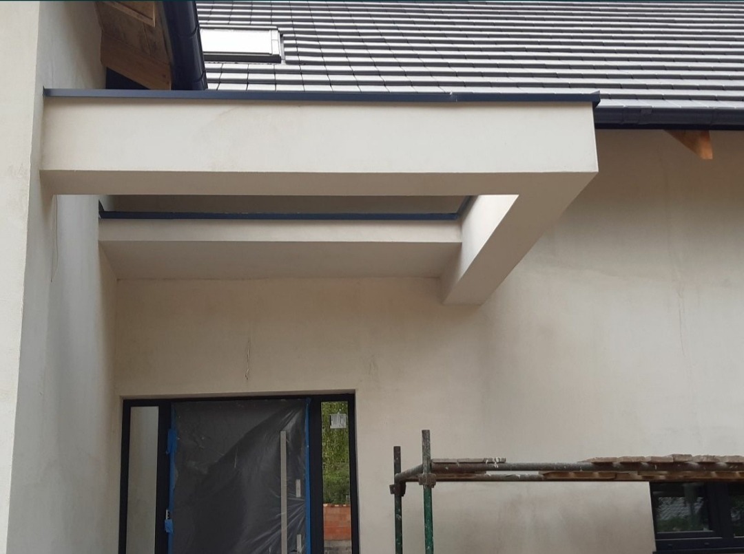 Elewacja domu w trakcie prac wykończeniowych, widoczne rusztowanie, zabezpieczone okno i świeżo otynkowana ściana z elementami architektonicznymi.