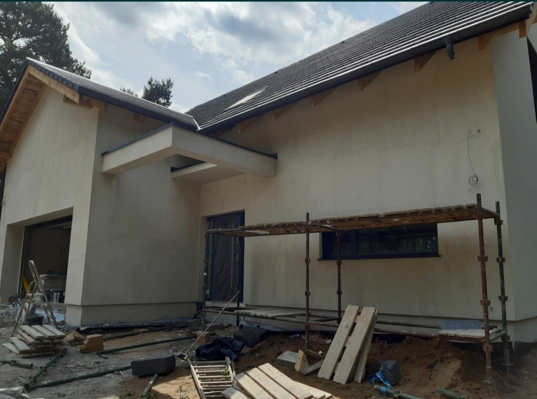 Dom jednorodzinny z elewacją w trakcie prac wykończeniowych, widoczne rusztowanie, materiały budowlane i lekko zachmurzone niebo.