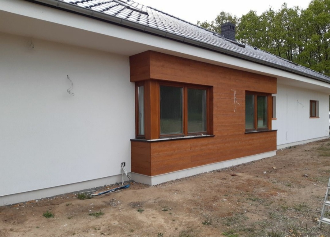 Fragment elewacji domu jednorodzinnego z drewnianą okładziną wokół okien, częściowo otynkowany na biało, z wystającymi kablami elektrycznymi i widoczną instalacją na gruncie.