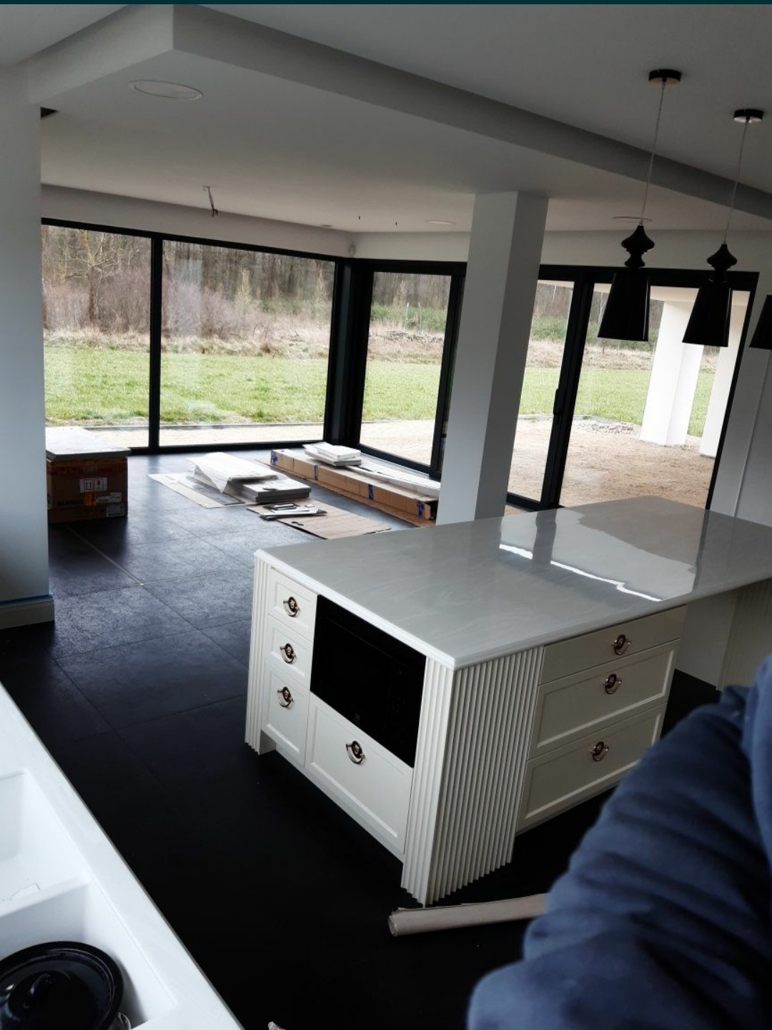 Wnętrze nowoczesnej kuchni z wyspą wykończoną białymi szafkami z frezowanymi detalami i marmurowym blatem, na czarnej podłodze, z dużymi oknami przesuwnymi z widokiem na zewnątrz, w trakcie prac...