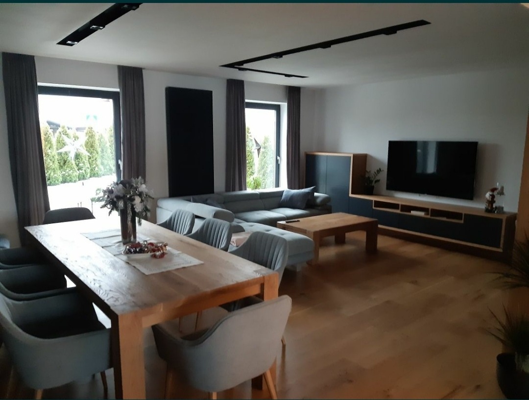 Jasne, przestronne wnętrze salonu z drewnianą podłogą, dużym narożnikiem, drewnianym stołem jadalnym z szarymi krzesłami i nowoczesną zabudową RTV z czarnym telewizorem.