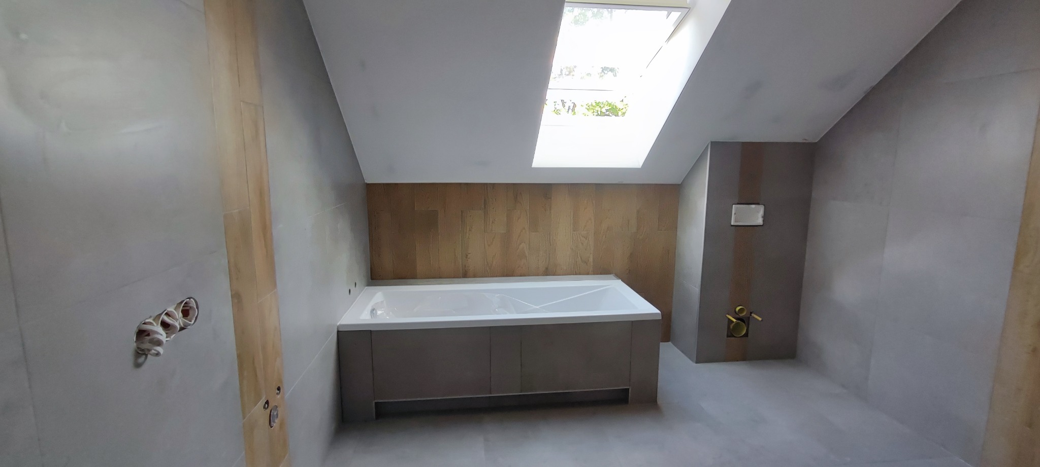 Wnętrze łazienki na poddaszu w trakcie wykańczania, z widoczną wanną w zabudowie, płytkami imitującymi drewno na ścianie, szarymi płytkami na podłodze i ścianach oraz oknem dachowym