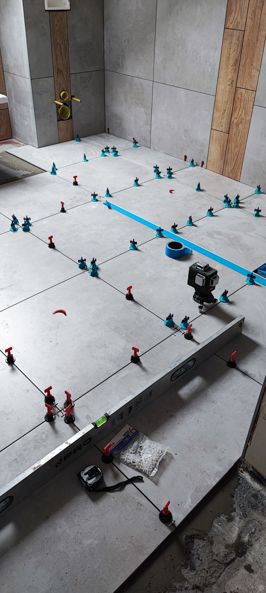 Szare płytki podłogowe w trakcie układania, z systemem poziomowania płytek (niebieskie i czerwone elementy), poziomica, miara zwijana i paczka krzyżyków dystansowych na podłodze, widoczne rury...