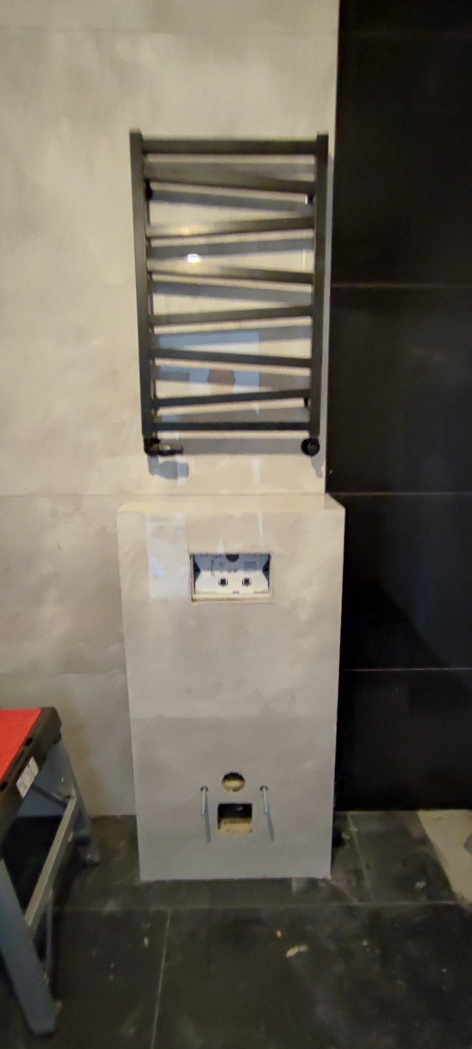 Montaż czarnego grzejnika drabinkowego na ścianie w łazience, widoczna instalacja podtynkowa i fragment drabiny malarskiej.