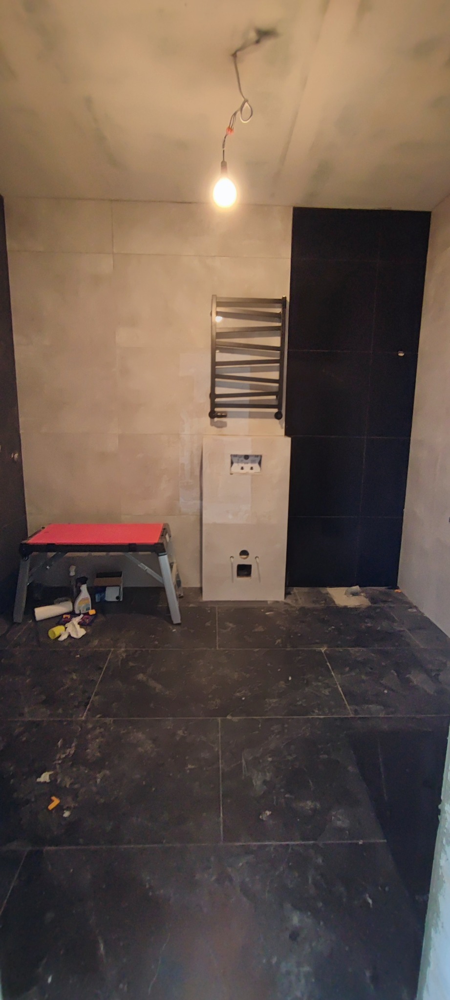 Wnętrze łazienki w trakcie prac: położona ciemna podłoga z płytek, częściowo wyłożone ściany jasnymi i czarnymi płytkami, zamontowany grzejnik drabinkowy, widoczny stelaż podtynkowy WC...