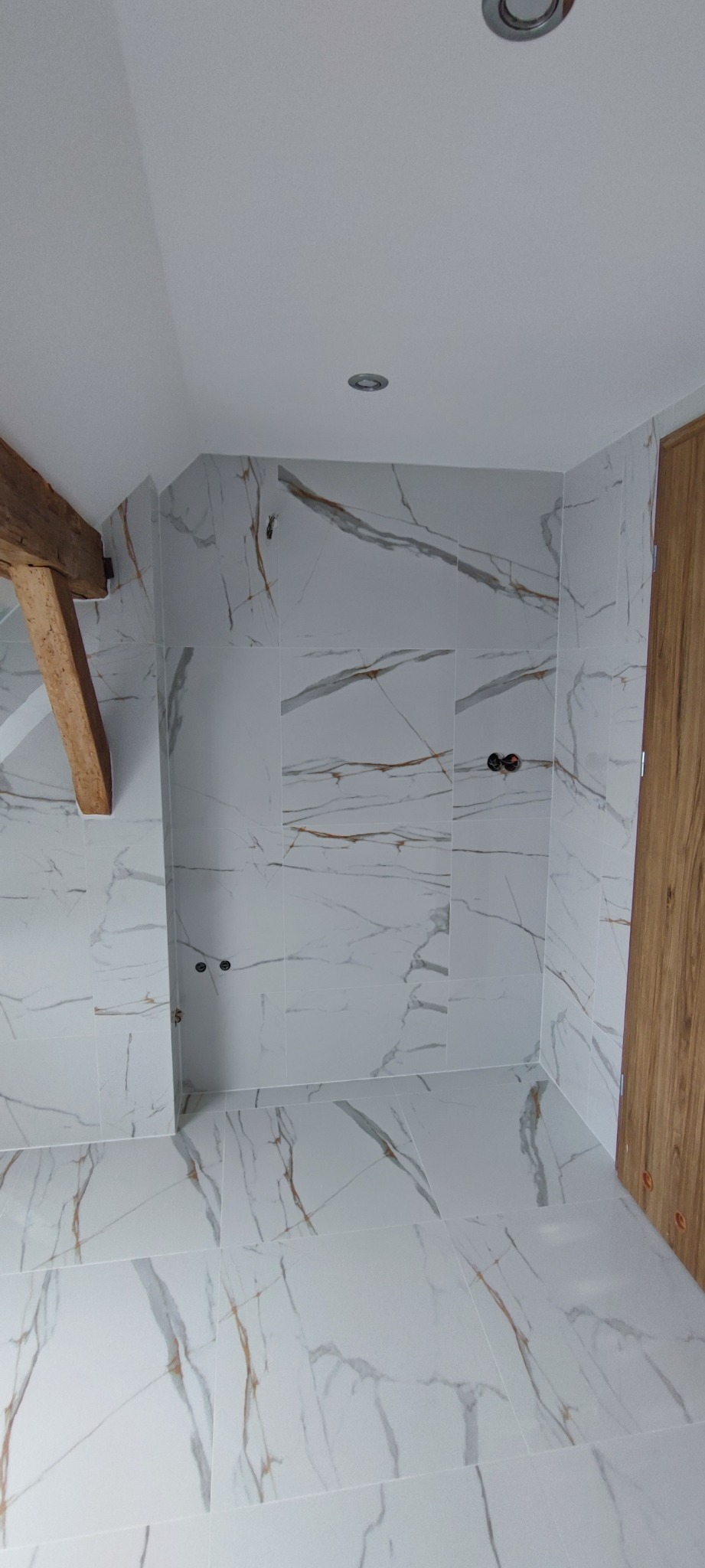 Wykończona łazienka z białymi płytkami imitującymi marmur, drewniana belka stropowa i drzwi, widoczne przyłącza wodne.