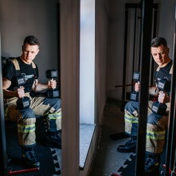 Trener Personalny Paweł Leśnicki - Mężczyzna w stroju strażackim siedzi na ławce i podnosi dwie czarne hantle 16kg w siłowni, widoczne elementy wyposażenia siłowni.
